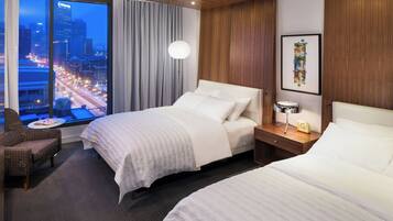 Habitación de hotel con dos camas, vista a la ciudad, un ventanal grande, una silla y una mesita de noche con lámpara y reloj.