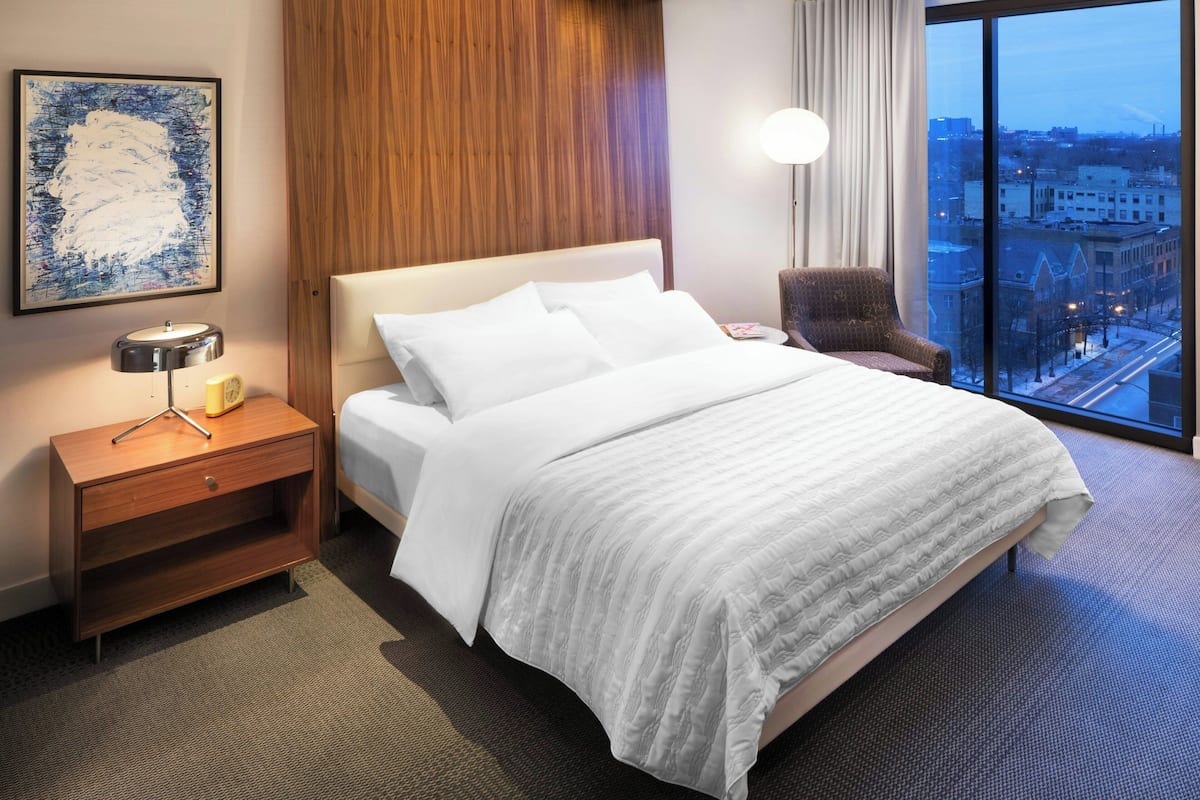 A hotel room with a large bed, a nightstand with a lamp, a chair, and a view of the city through a large window.