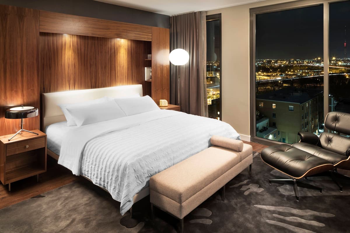 A modern hotel room with a large bed, a bench, a leather chair, a nightstand, and a view of a cityscape at night.