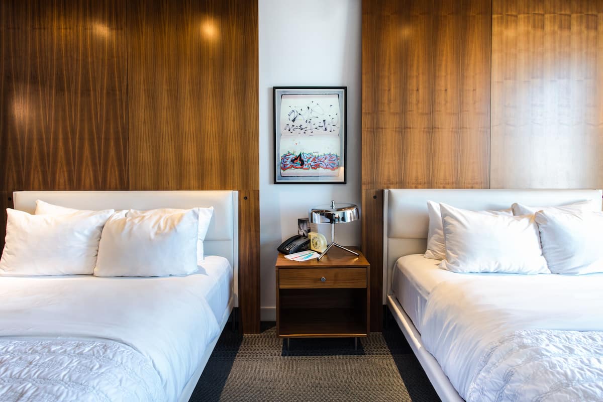 A hotel room with two beds, a wooden wall, a framed artwork, a nightstand with a lamp, and a small table.