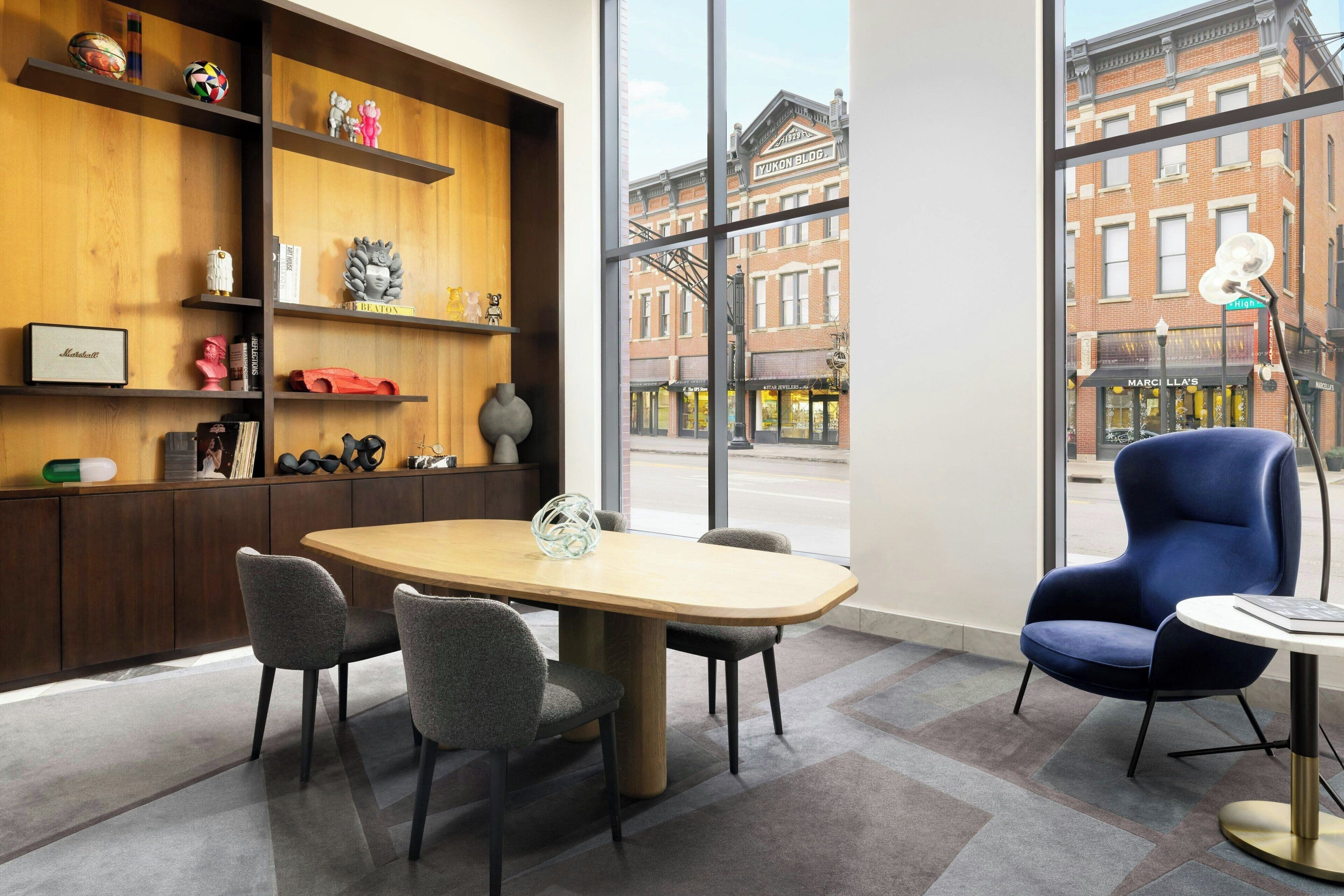 A modern room with a wooden table, four chairs, a blue armchair, and a view of a brick building outside.