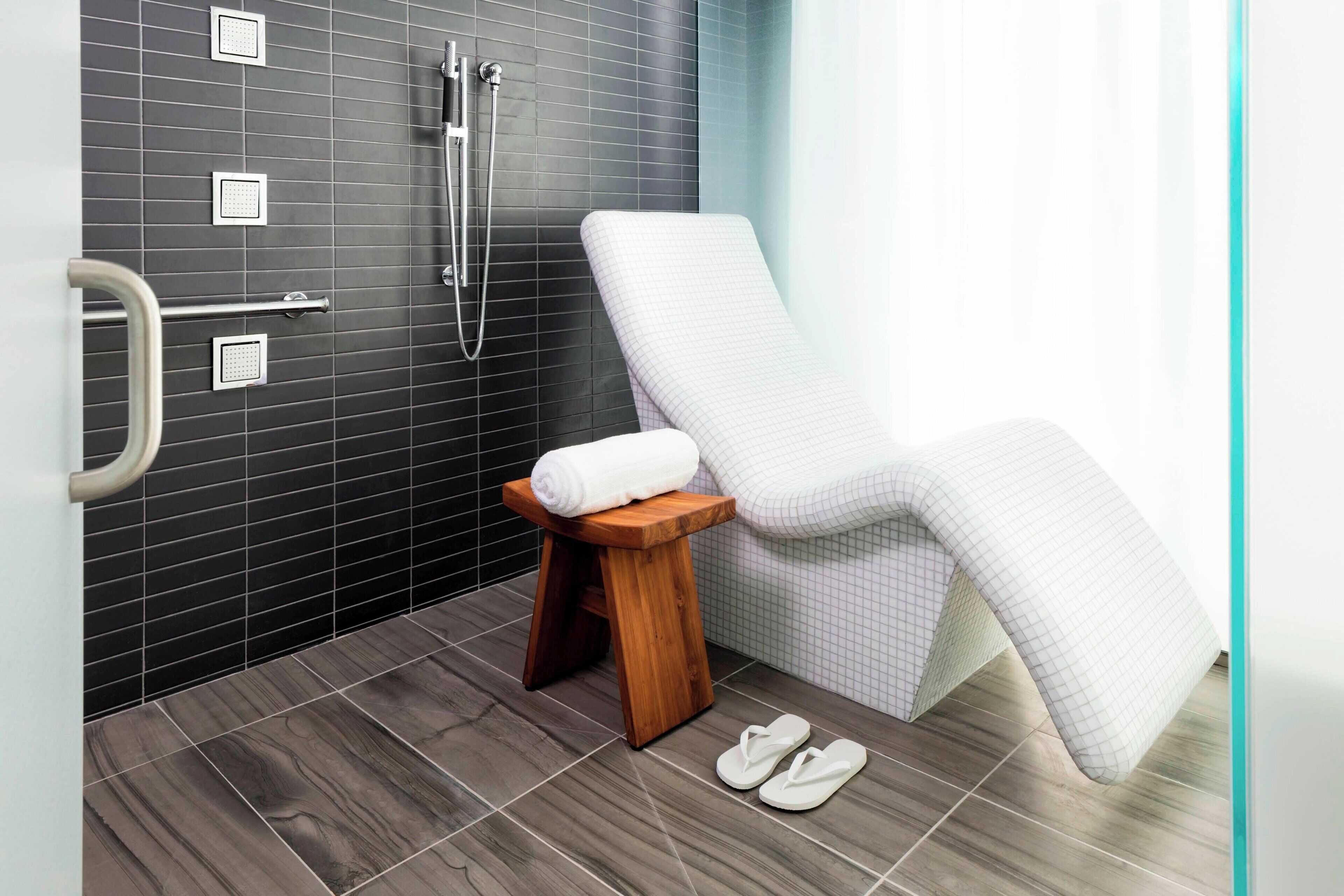A modern bathroom with a shower, a towel on a stool, and a pair of slippers on the floor.
