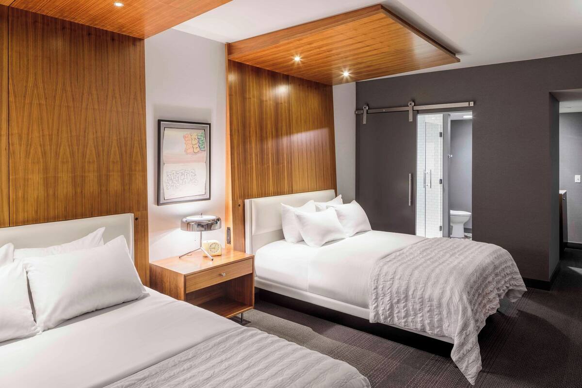 A hotel room with two beds, wooden paneling, a framed picture, a bedside table with a lamp, and a bathroom visible through an open door.