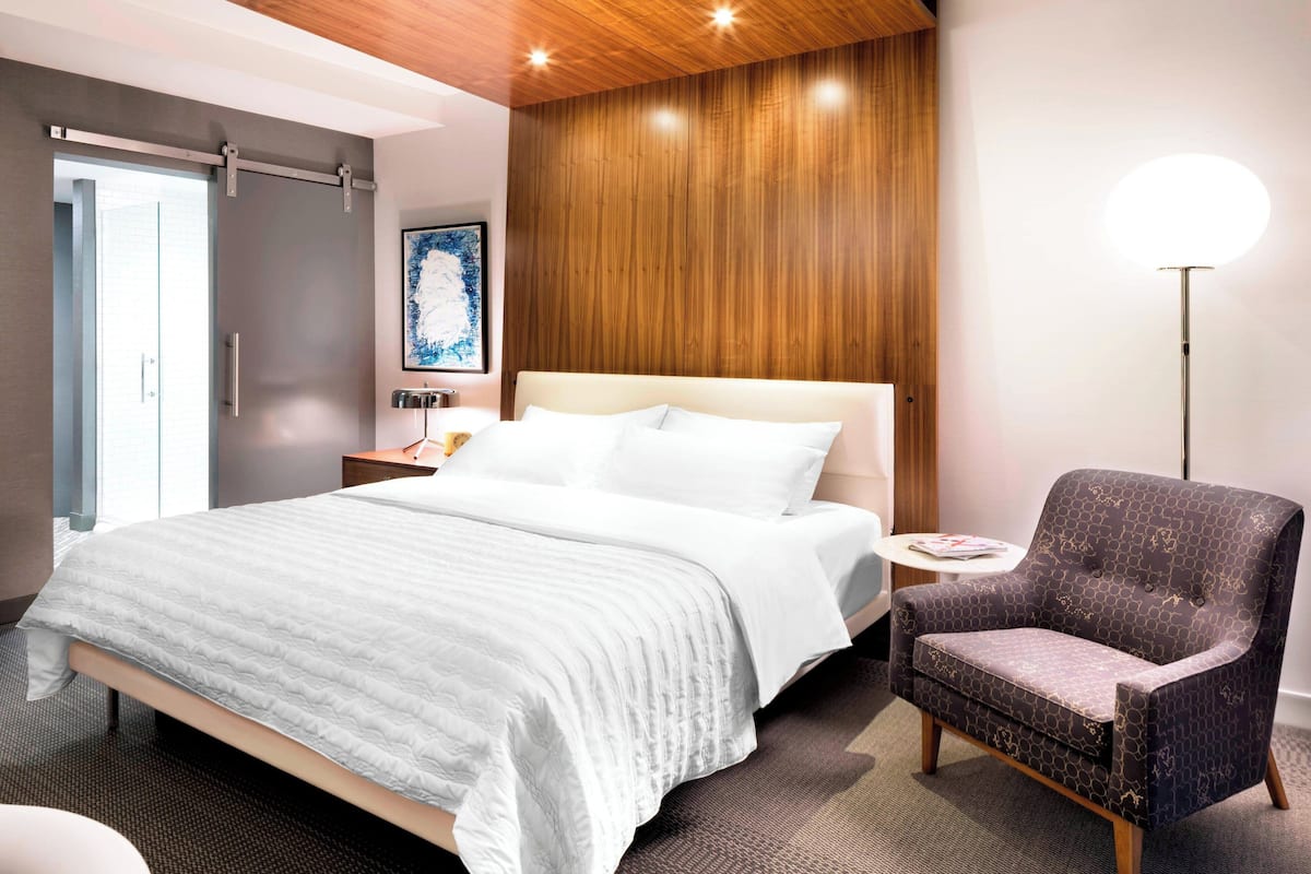 A modern hotel room with a large bed, a wooden accent wall, a standing lamp, and a small side table.