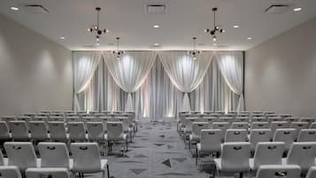 Un salón de conferencias con filas de sillas blancas, una alfombra estampada y un amplio ventanal con cortinas translúcidas.