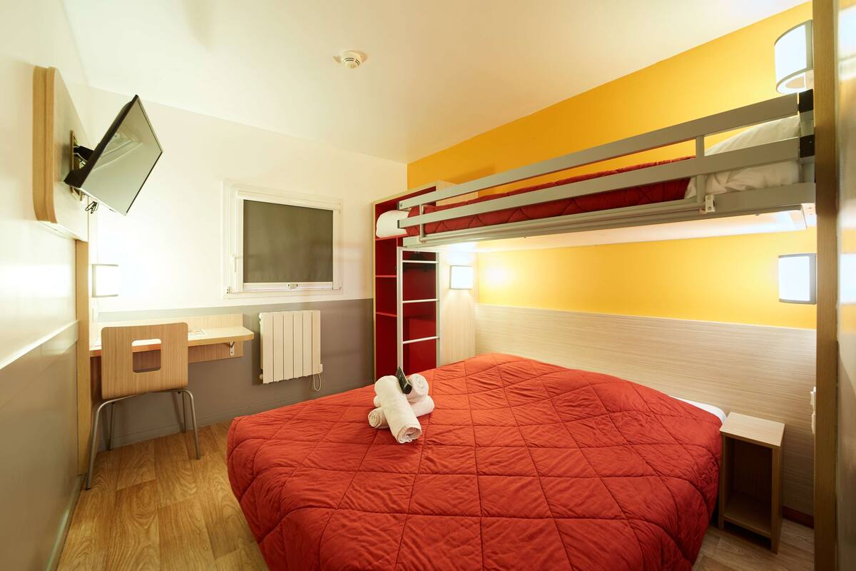 Een slaapkamer met stapelbed, een rode sprei, een bureau met stoel, een ladder en een aan de muur bevestigde televisie.
