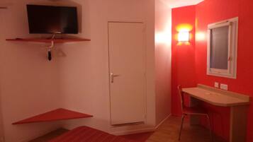 Een kamer met een witte deur, een rode muur, een klein tafeltje, een stoel en een tv aan de muur.