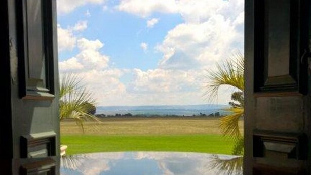 Vista a través de una ventana con marco decorativo, que refleja un paisaje con cielo despejado, nubes y follaje.