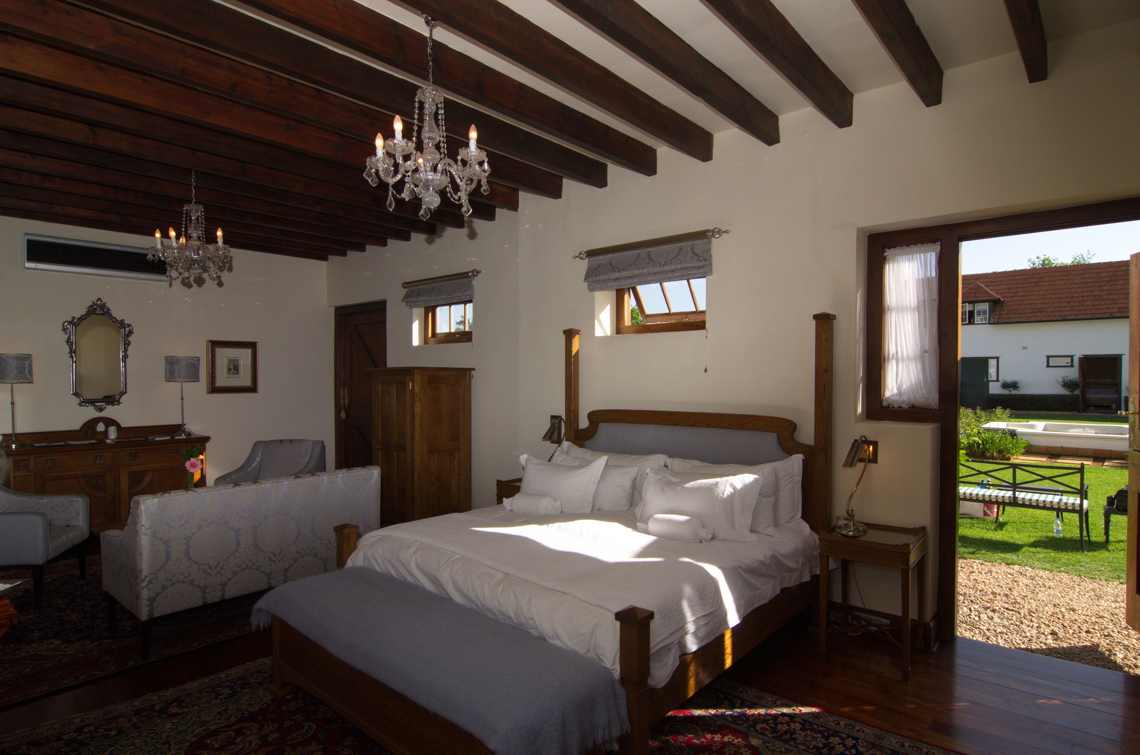 A bedroom with a large bed, two bedside tables, a wooden dresser, a chandelier, and a view of the outside.
