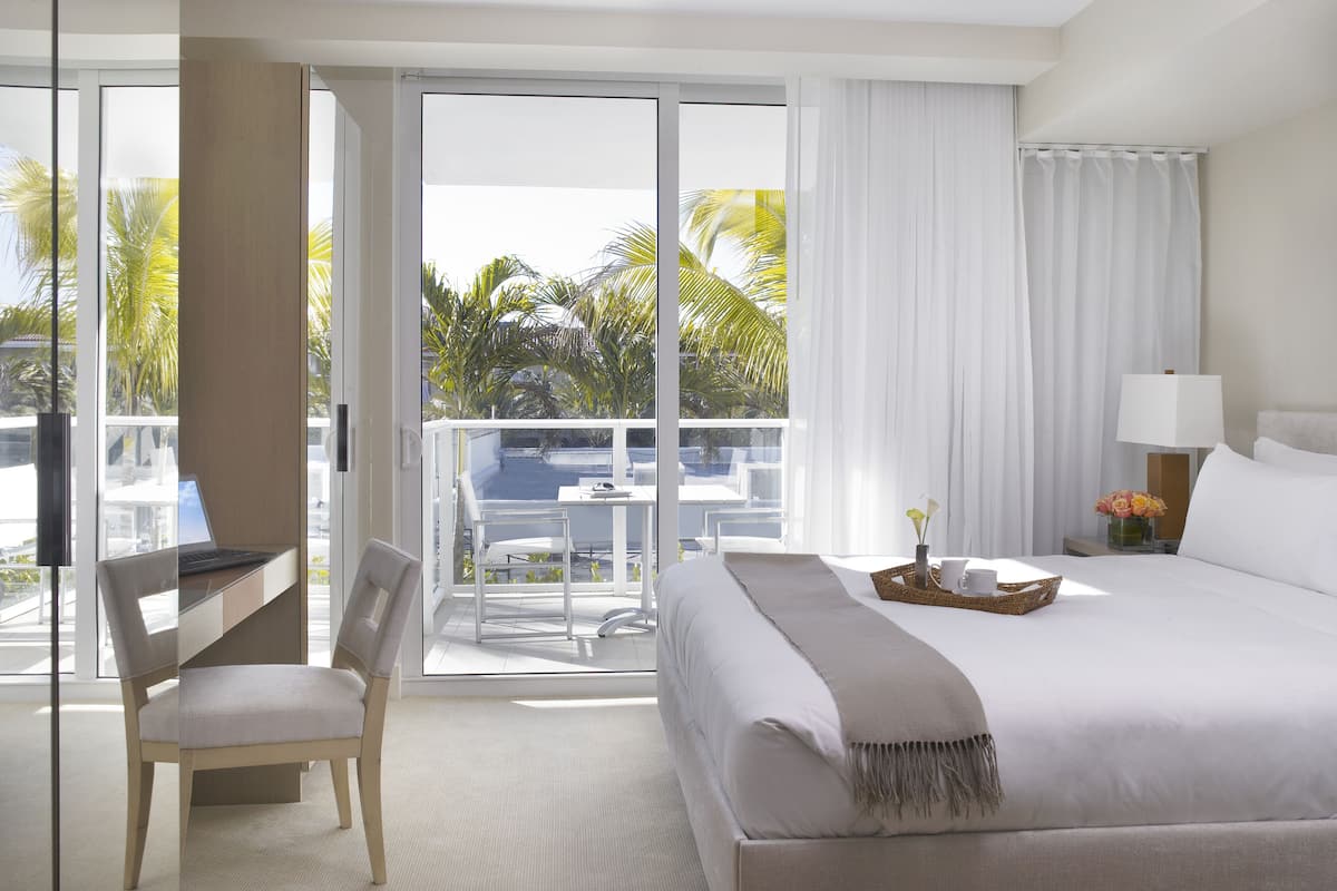 Une chambre d’hôtel avec un lit, un bureau, une chaise, une lampe, un vase de fleurs et une vue sur les palmiers, visible à travers de grandes fenêtres.