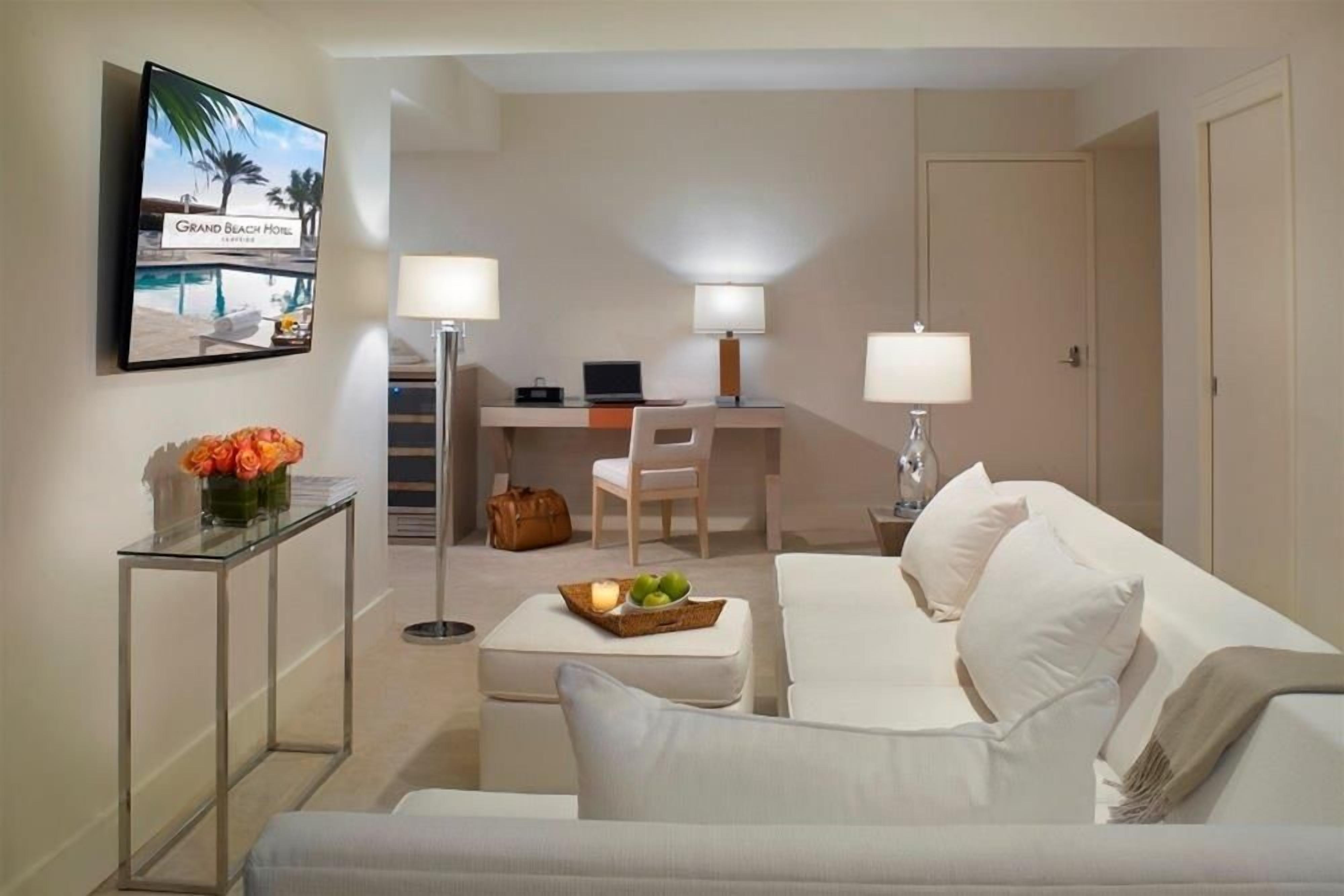 A hotel room with a bed, a desk with a laptop, a chair, and a TV displaying the hotel name.