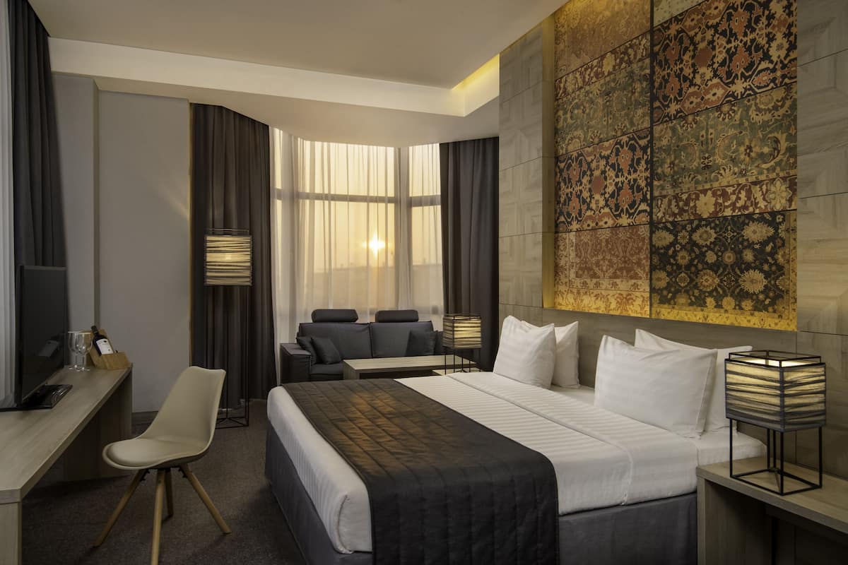 A hotel room with a bed, a desk, a chair, a sofa, and a large window with curtains.