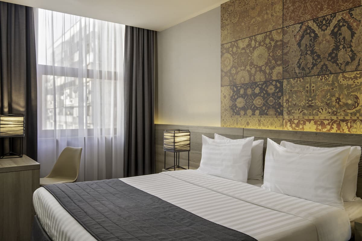 A hotel room with a large bed, a desk, a chair, a lamp, and a decorative wall panel.