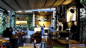 Un café en plein air avec des tables et des chaises en bois, un comptoir où est affiché le menu, et de la verdure.