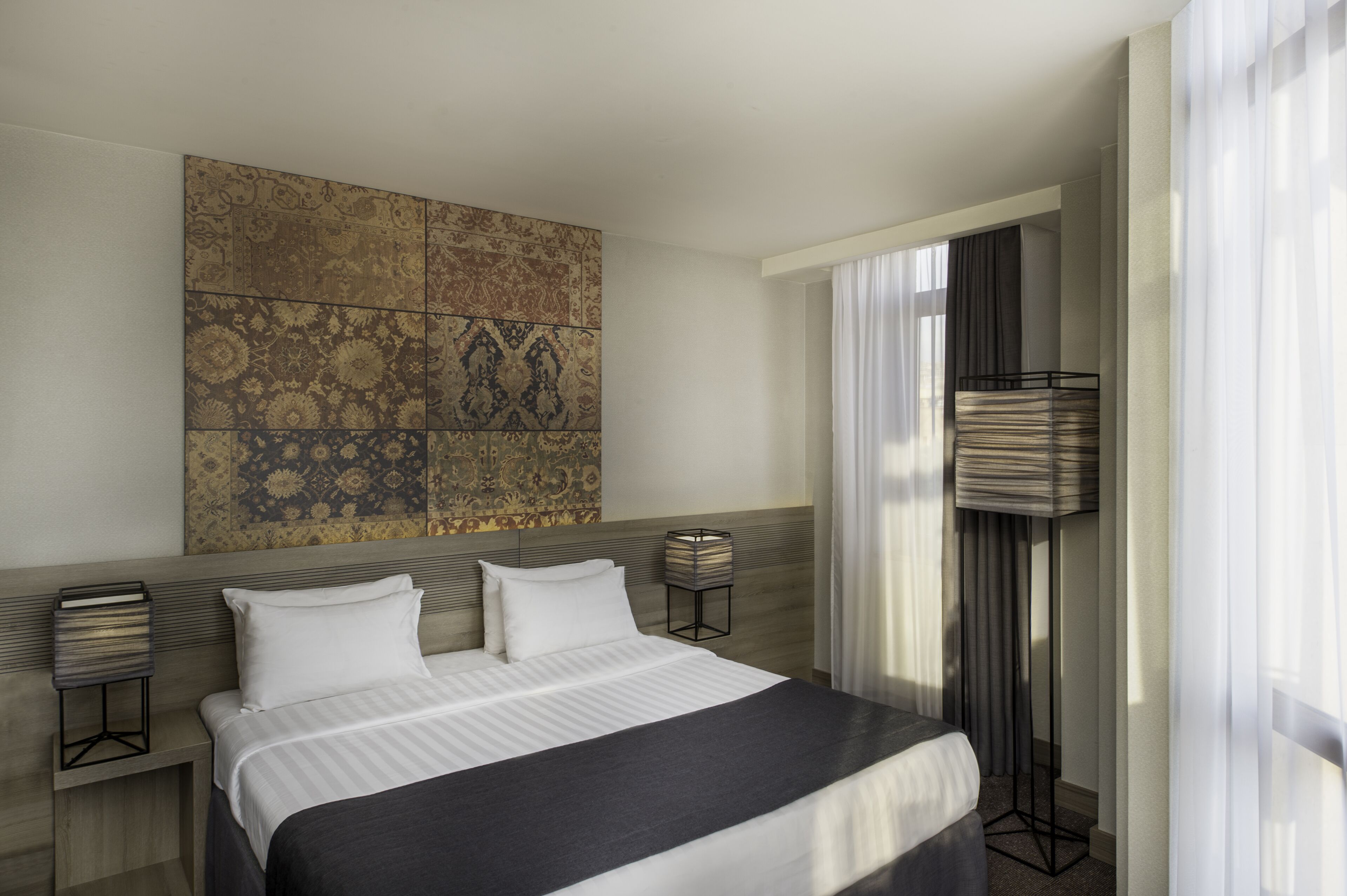 A modern hotel room with a large bed, a decorative wall panel, a floor lamp, and a view of the outdoors.