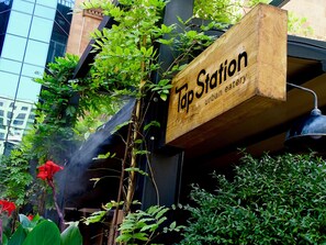A wooden sign with the text "Tap Station urban eatery" hanging outdoors, surrounded by greenery and a building in the background. - Republica Hotel Yerevan (Yerevan)
