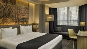 Une chambre d’hôtel dotée d’un grand lit, d’un canapé, d’un bureau et offrant une vue sur la ville grâce à de grandes fenêtres.