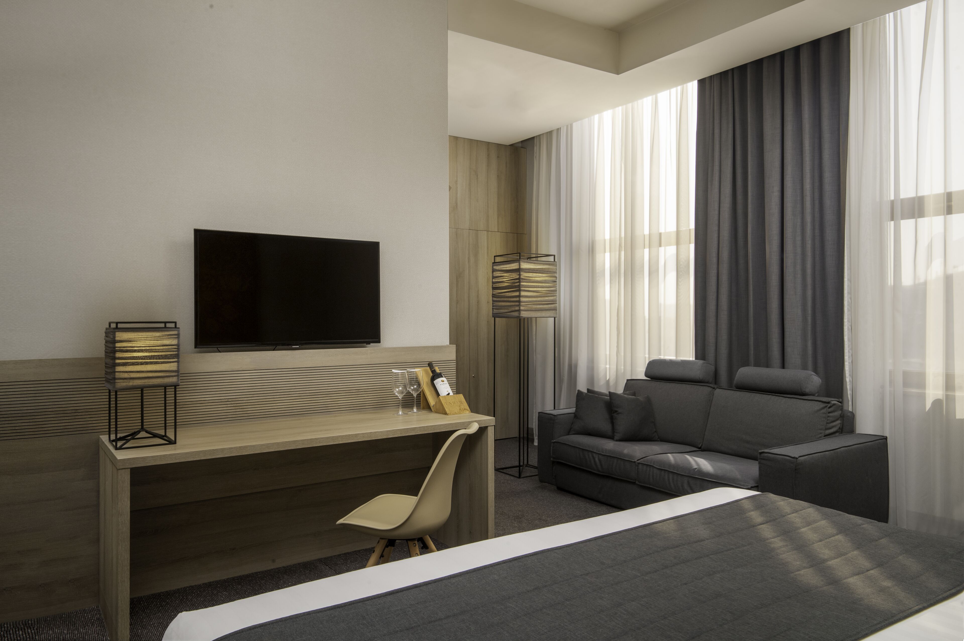 A modern hotel room with a bed, a desk with a chair, a sofa, a TV mounted on the wall, and a floor lamp.