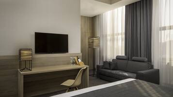 A modern hotel room with a bed, a desk with a chair, a sofa, a TV mounted on the wall, and a floor lamp.