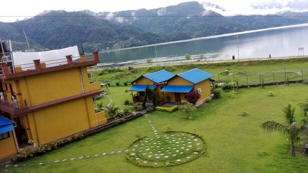 A yellow building with a blue roof, a circular garden with stepping stones, and a view of a lake and mountains in the background. - Hotel Lakefront (Pokhara)