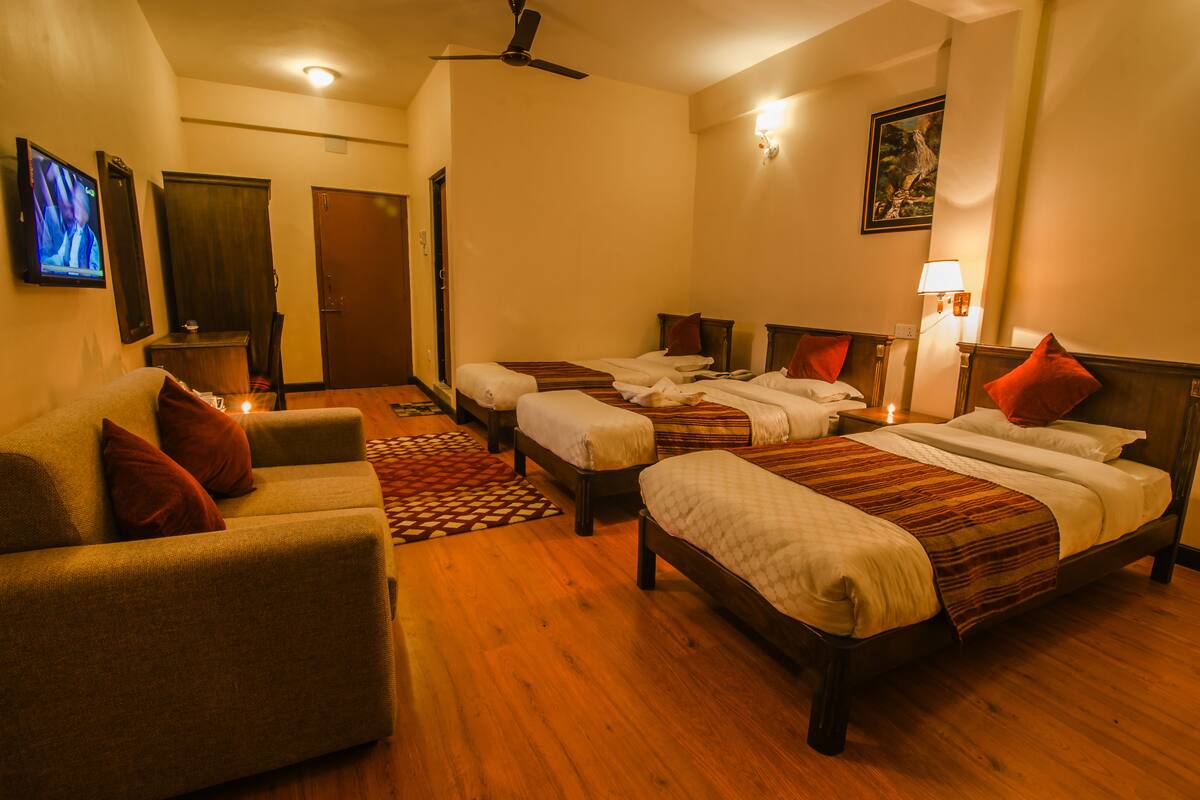 A hotel room with a sofa, two single beds, a TV, a ceiling fan, and a painting on the wall.