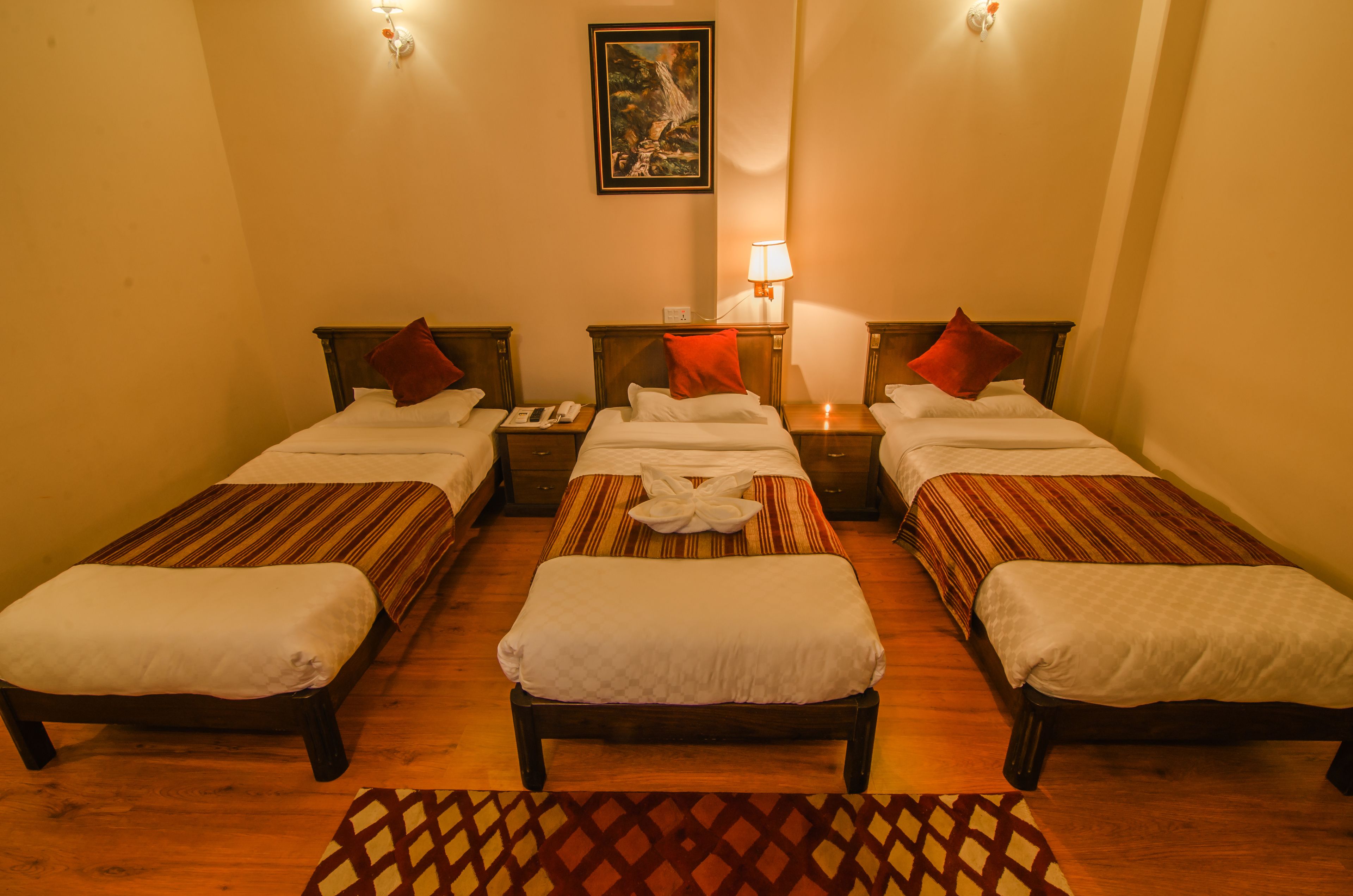 A hotel room with three single beds, a nightstand with a lamp, and a painting on the wall.