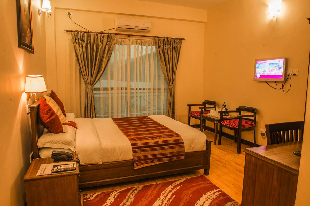 A hotel room with a bed, bedside table, lamp, TV, and a small dining area with chairs.
