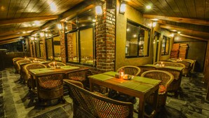 A restaurant with wicker chairs and tables, brick pillars, and large windows. - Hotel Lakefront (Pokhara)