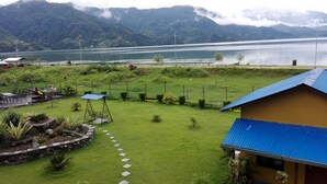 A lush green lawn with a swing set, a small house with a blue roof, and a view of a lake and mountains in the background. - Hotel Lakefront (Pokhara)