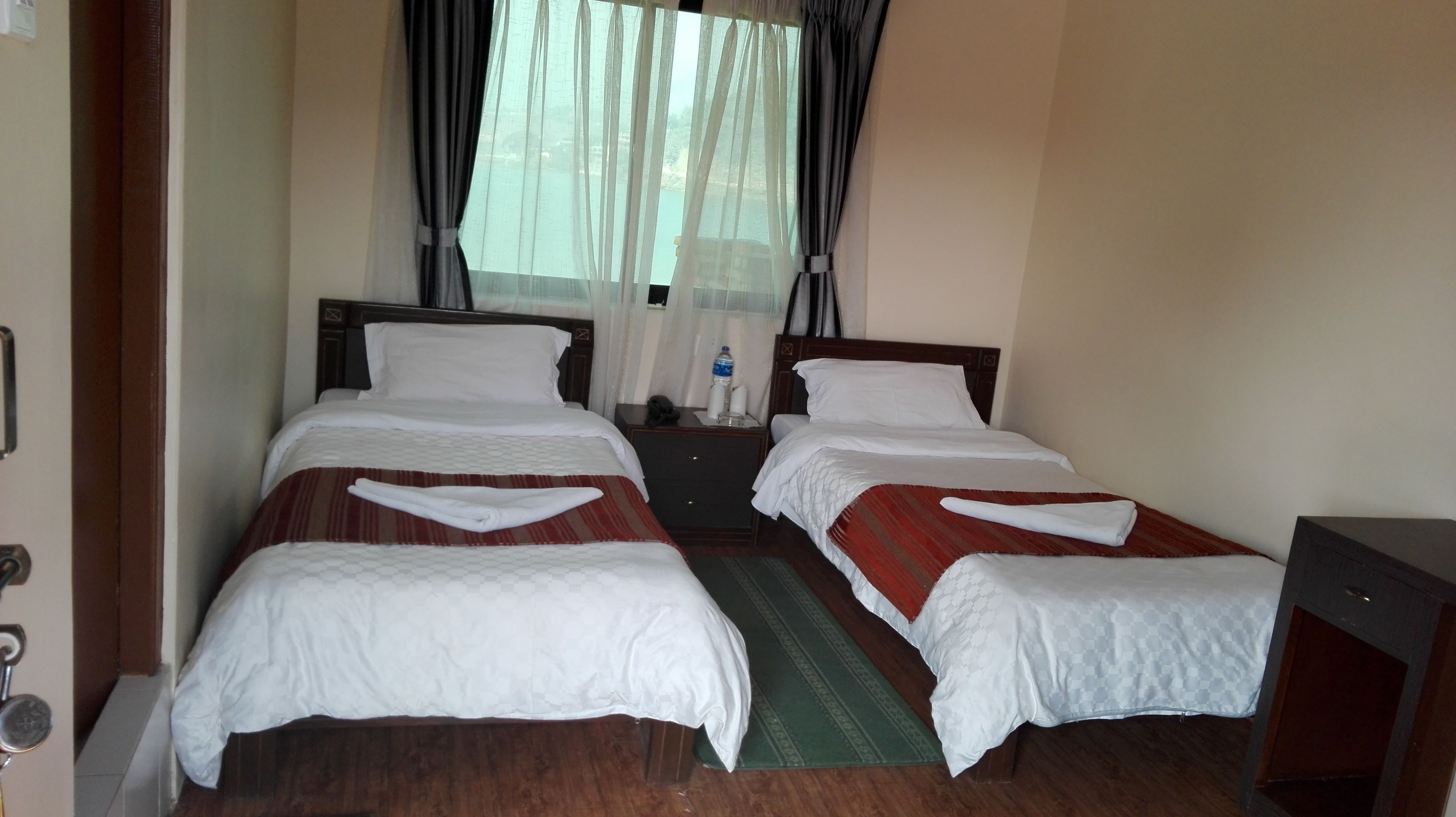 A hotel room with two beds, a nightstand, a small table, and a window with sheer curtains.