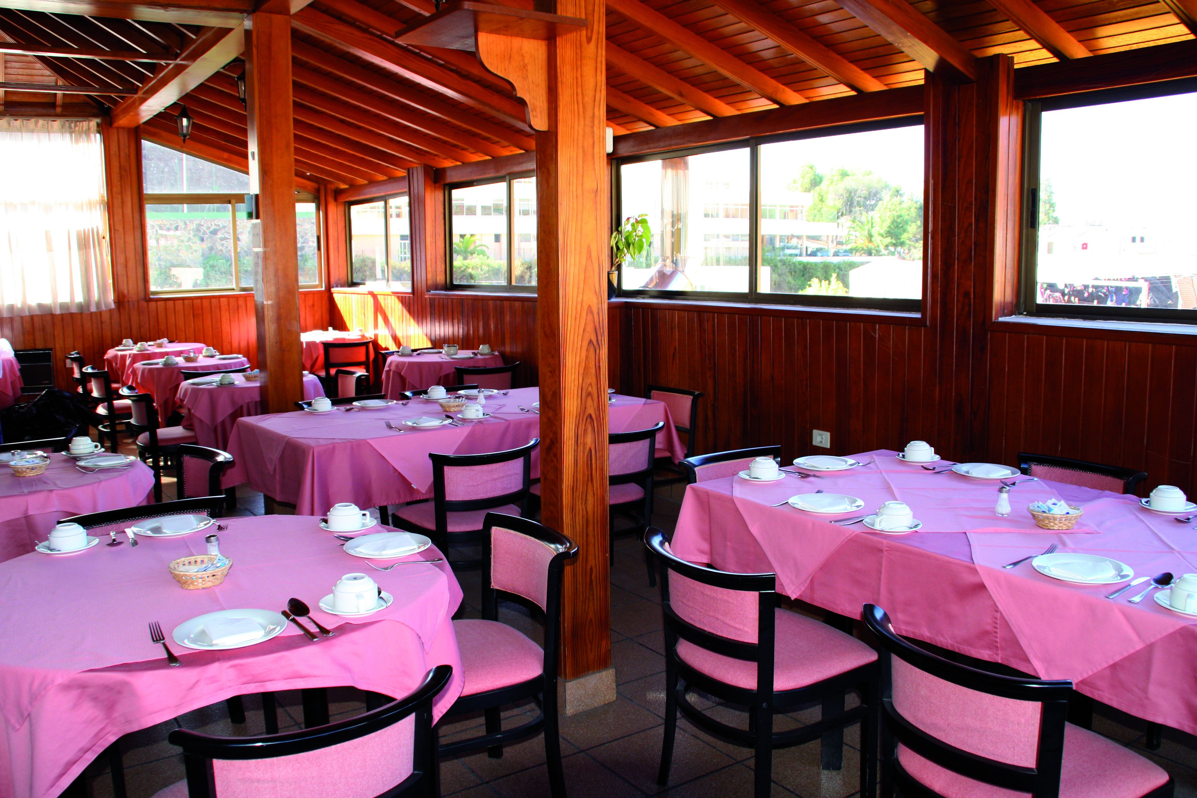 A restaurant with multiple tables covered in pink tablecloths, wooden beams on the ceiling, and large windows offering a view of the outdoors.