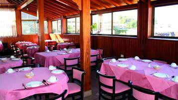 A restaurant with multiple tables covered in pink tablecloths, wooden beams on the ceiling, and large windows offering a view of the outdoors.