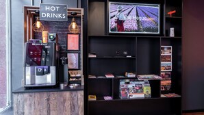 Máquina de café, placa indicando bebidas quentes, televisão exibindo 'The Hague' e uma estante com diversos objetos.