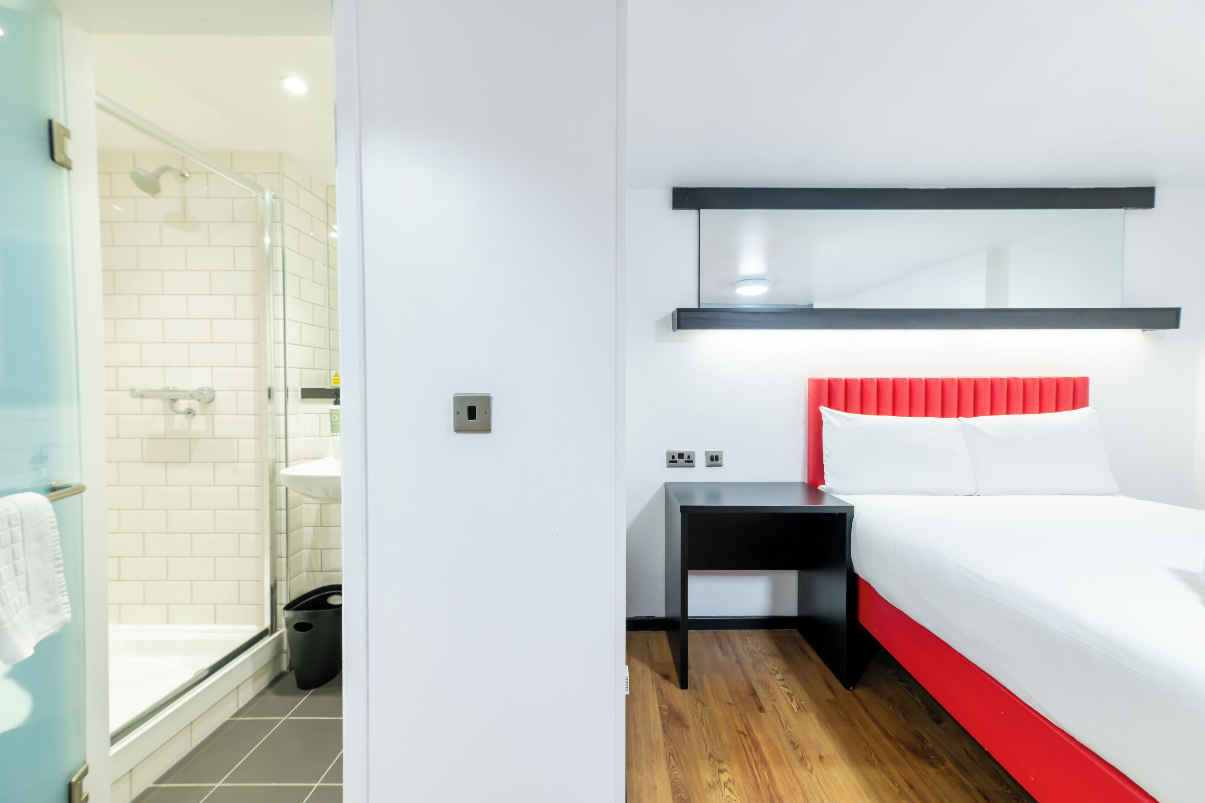 A hotel room with a shower, a bedside table, a bed with a red headboard, and a mirror above the bed.