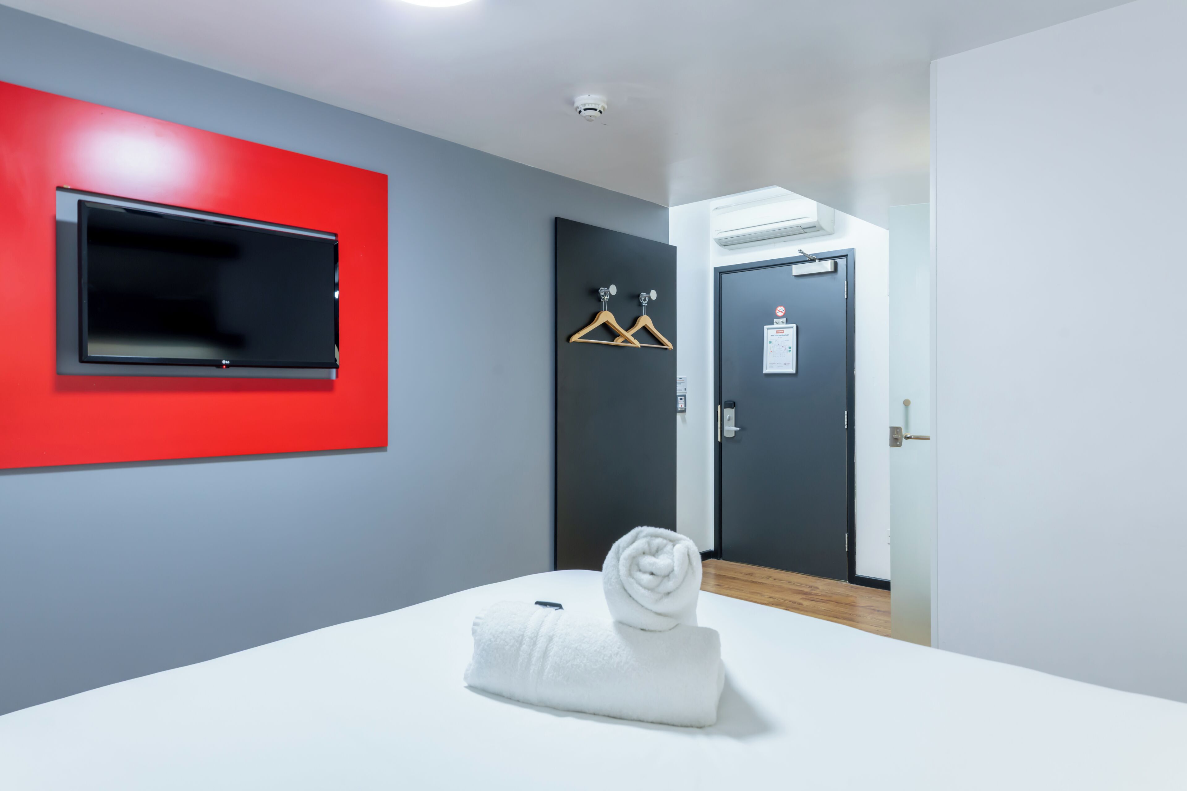 A hotel room with a flat-screen TV mounted on a red wall, a towel on the bed, a door with a coat hanger, and an air conditioning unit on the wall.