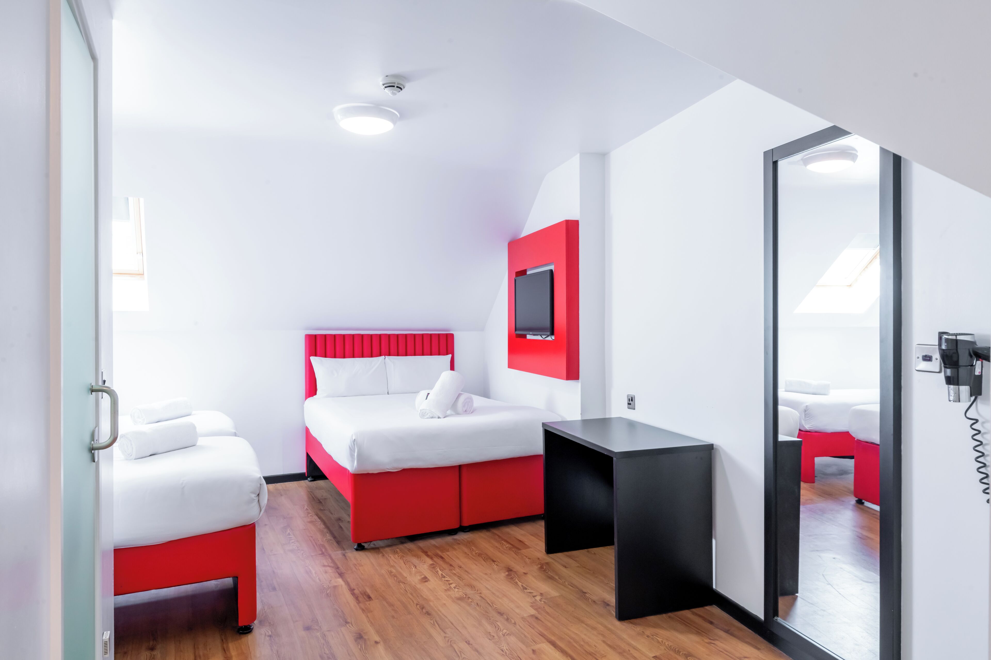 Moderni hotellihuone, jossa on punavalkoinen sänky, pieni musta pöytä ja seinälle asennettu televisio.