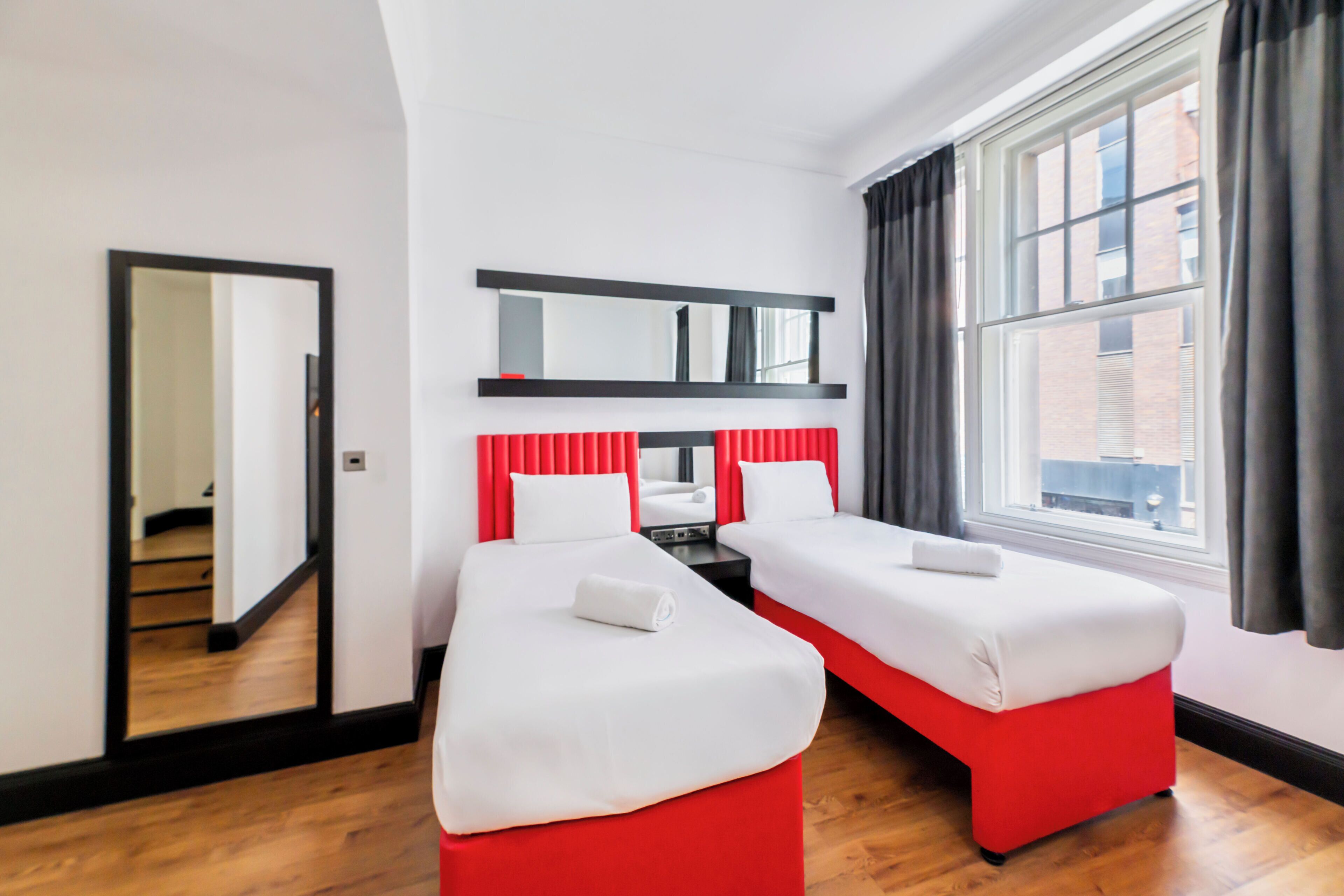 A hotel room with two single beds, a large mirror, and a window with curtains.