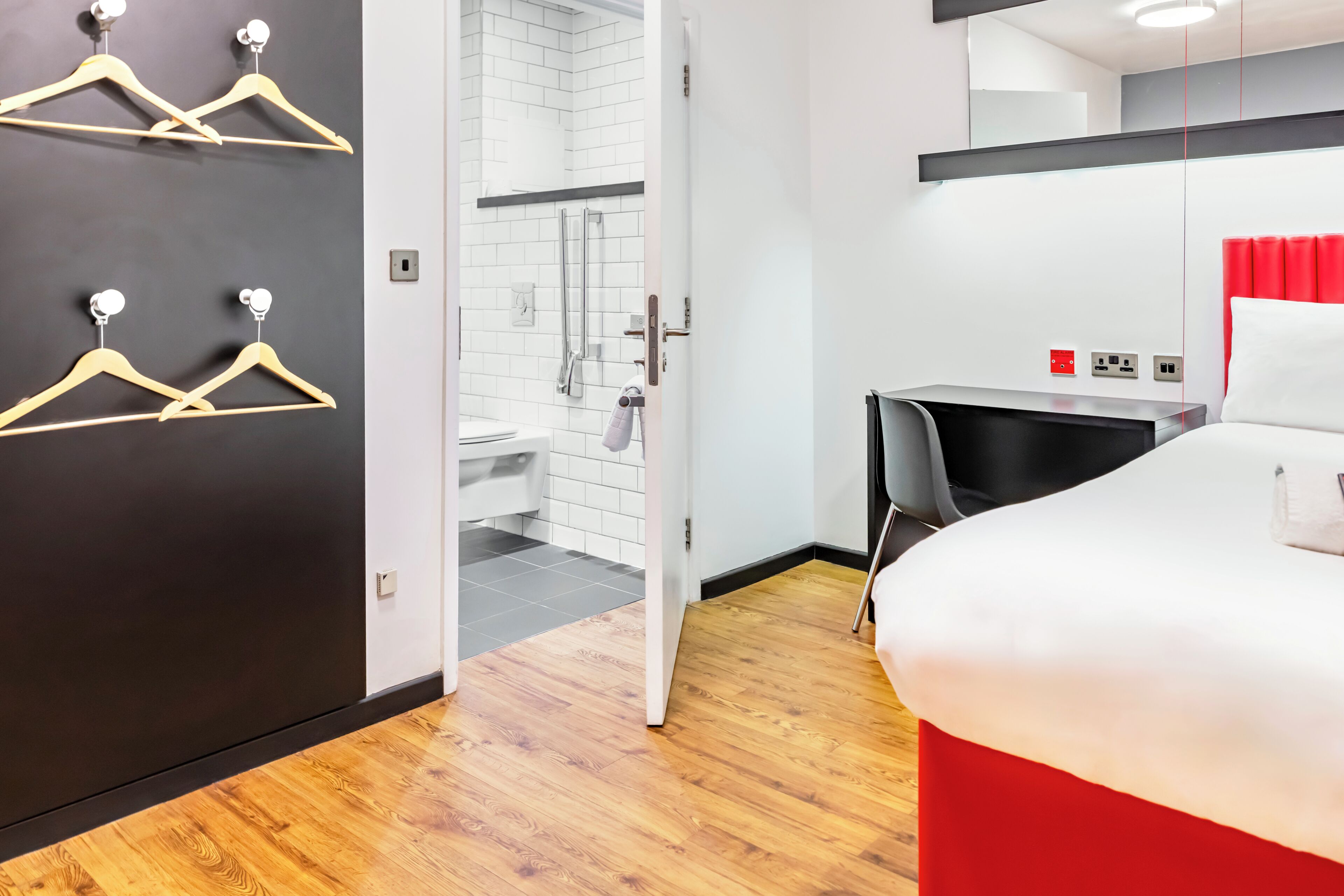 A hotel room with a bed, a desk, a chair, a wardrobe, and a bathroom visible through an open door.