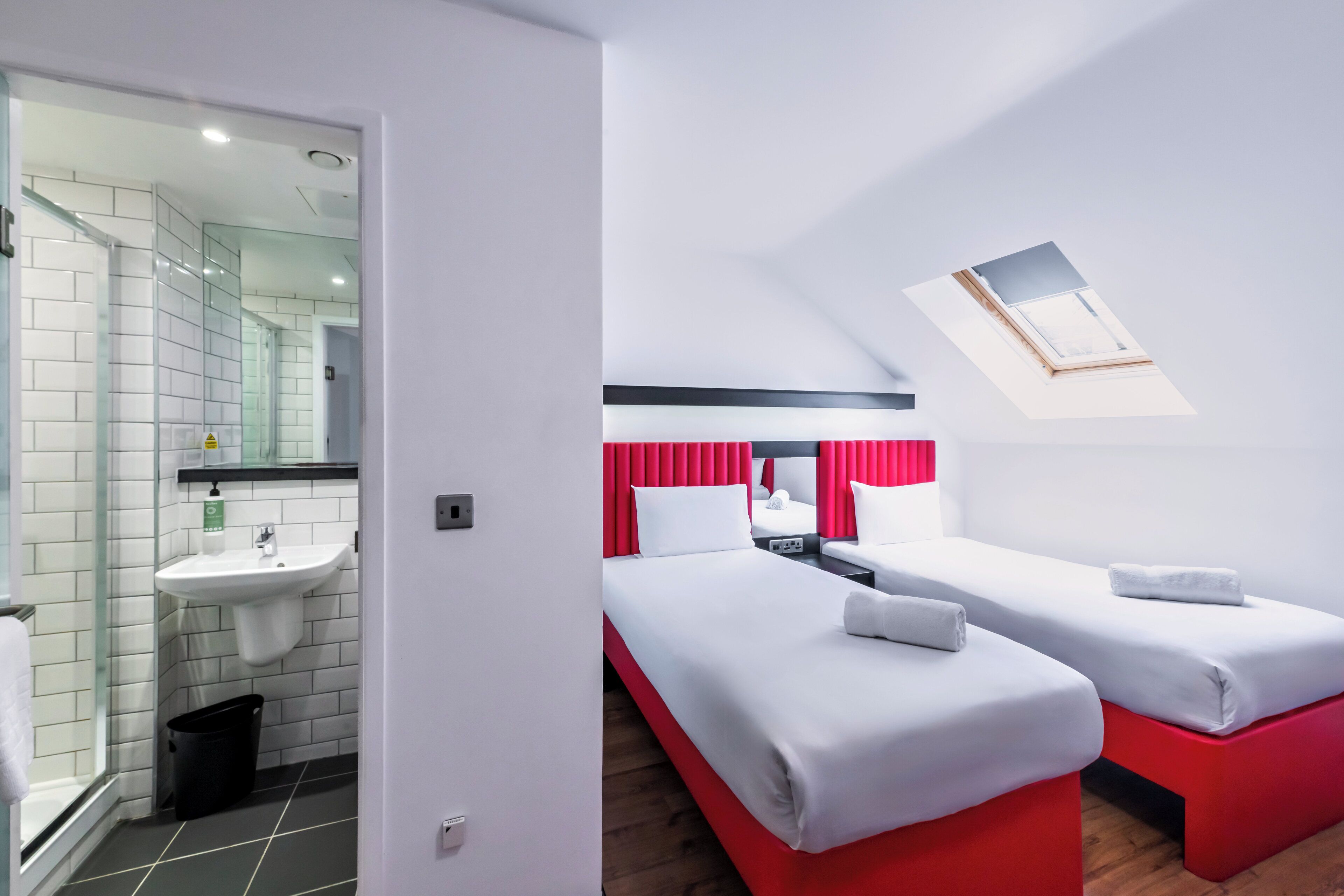 A hotel room with a bathroom, a skylight, and two beds with red bases.
