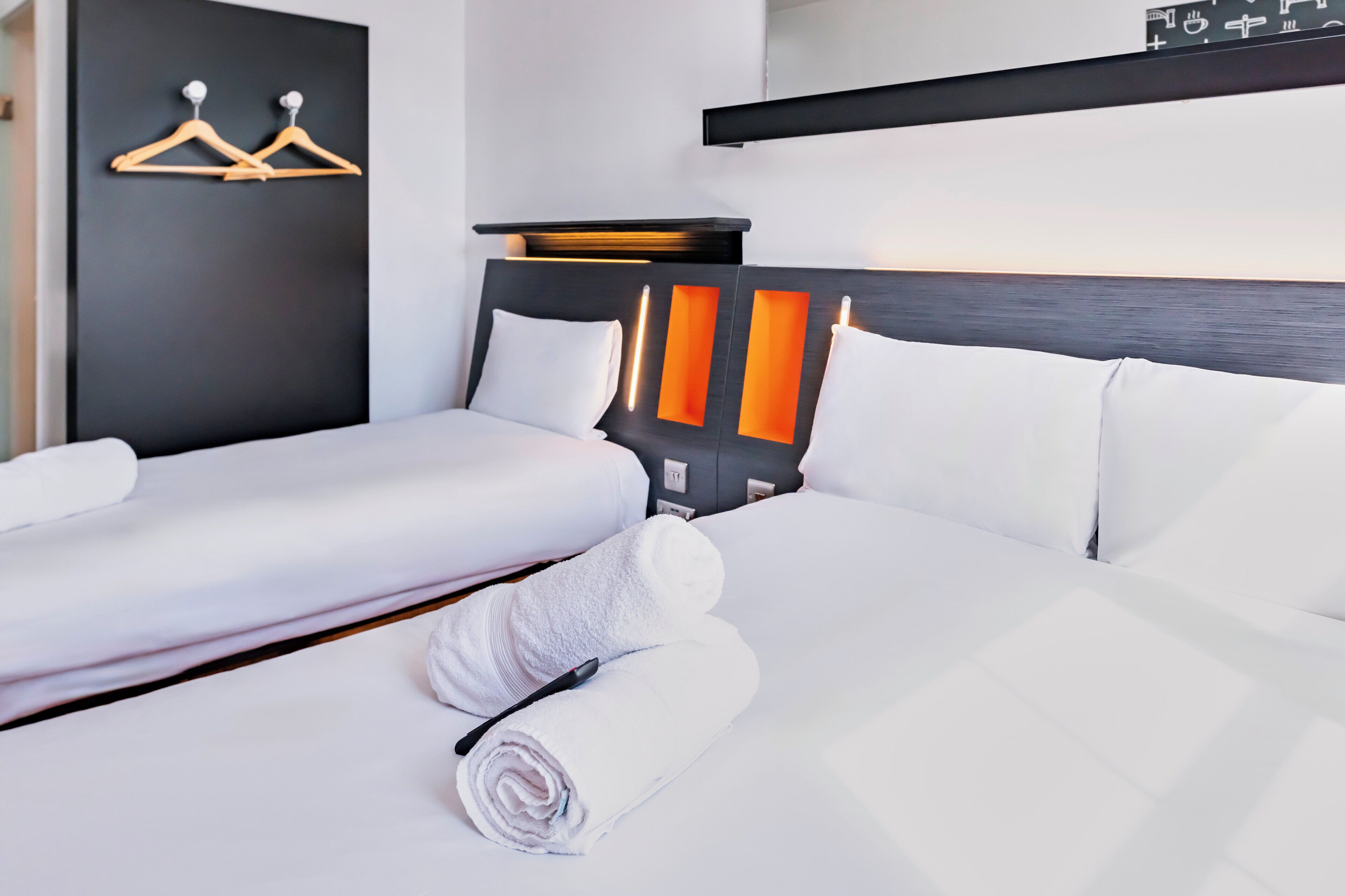 Ett hotellrum med två sängar, vita sängkläder, en svart sänggavel med orange detaljer och en väggmonterad handduksställning.
