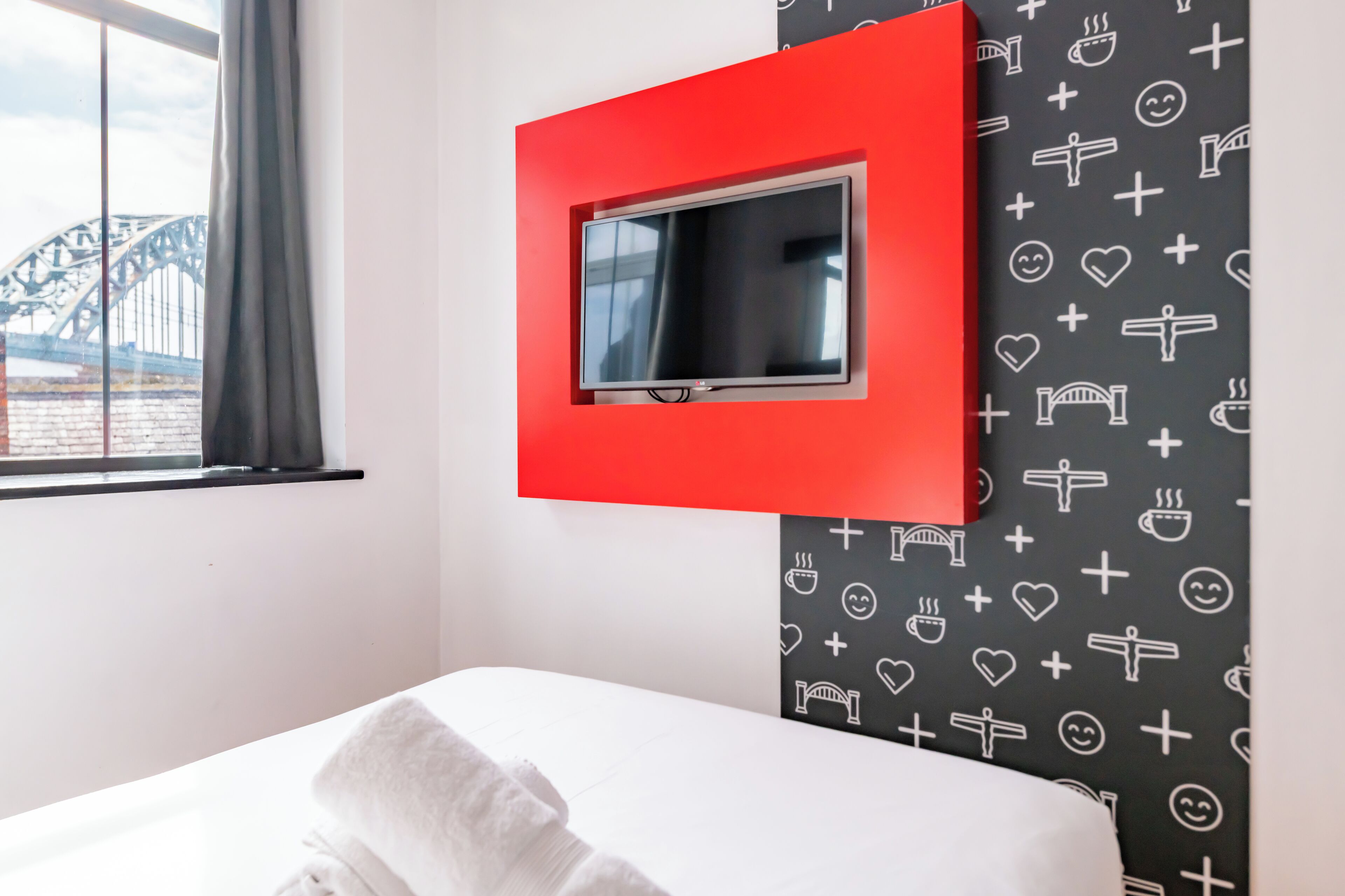 A hotel room with a bed, a window with a view of a bridge, and a TV mounted on a red frame.