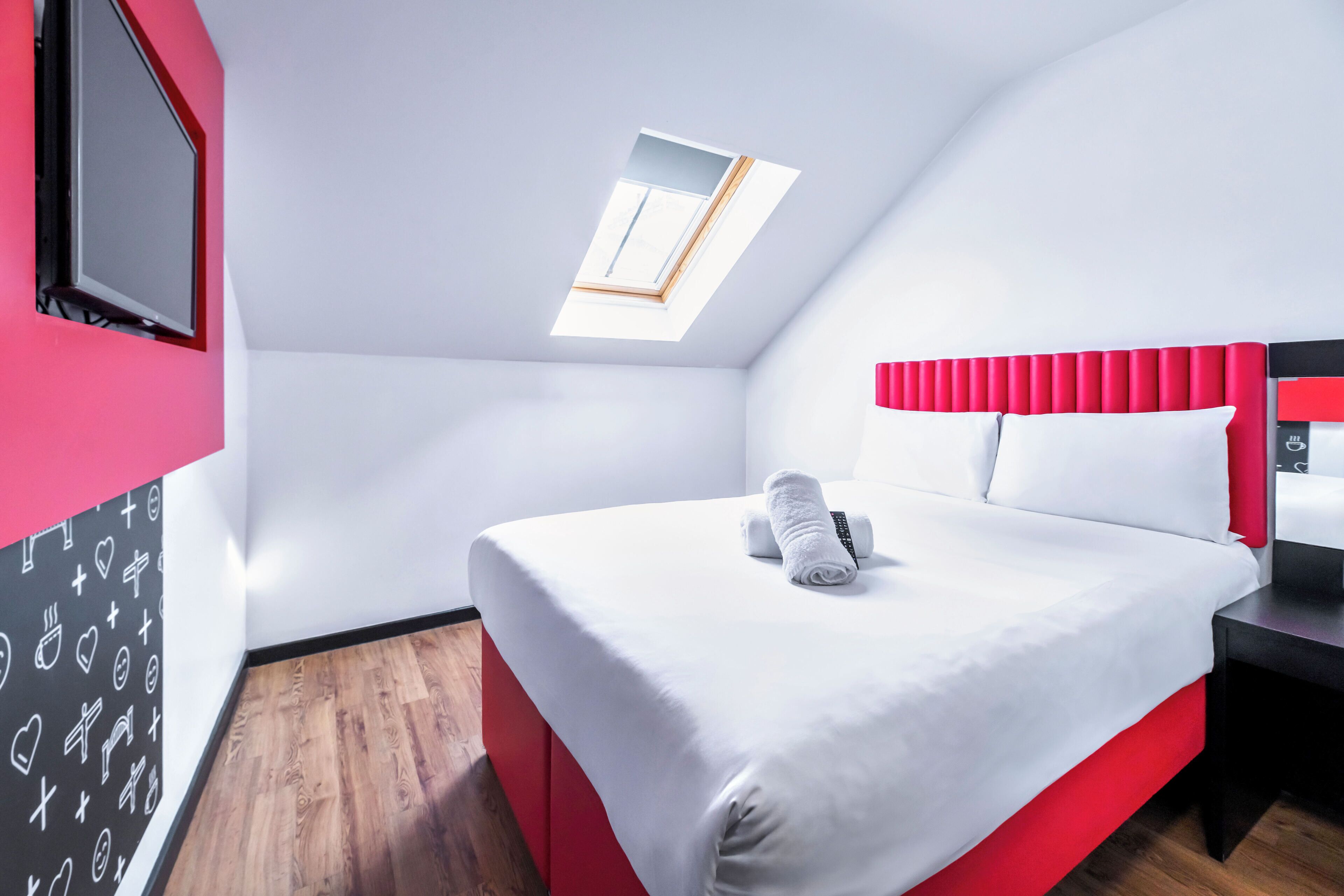 A modern hotel room with a bed, a TV, a skylight, and a red accent wall.