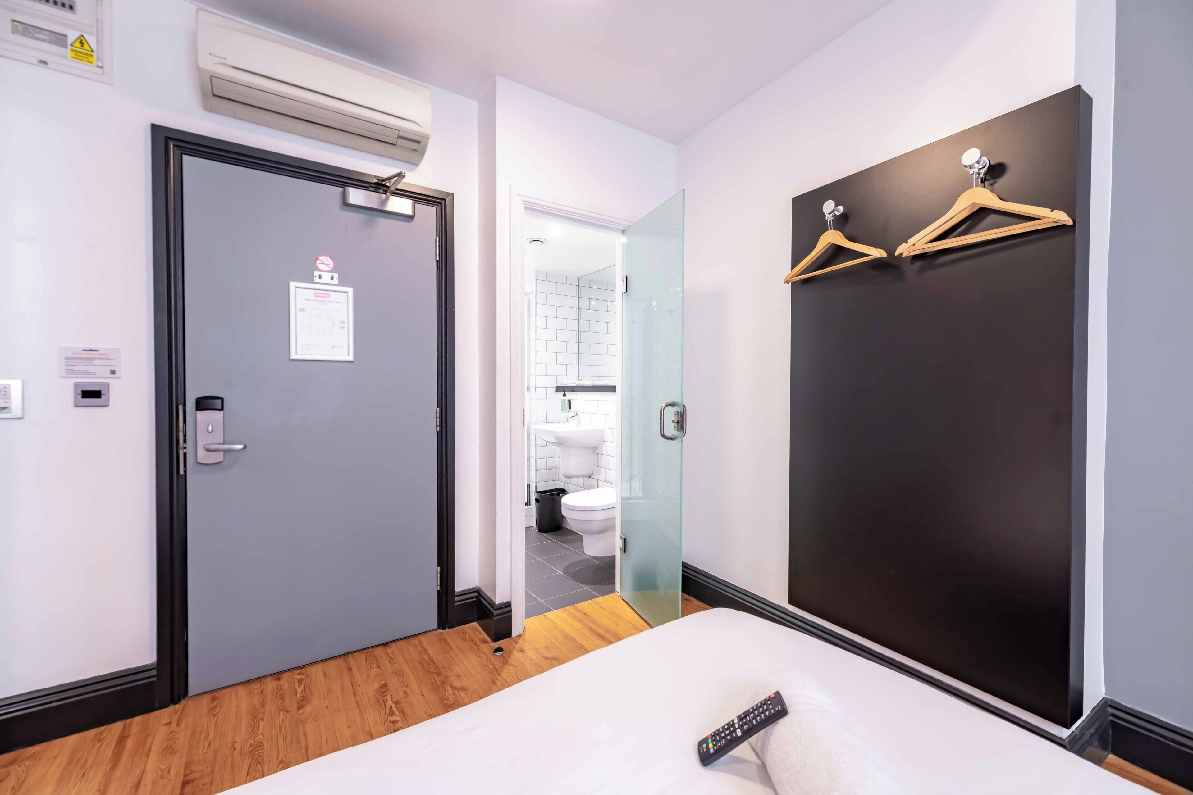Une chambre d’hôtel comprenant un lit, une porte, un climatiseur fixé au mur, un luminaire fixé au mur et un porte-manteau.