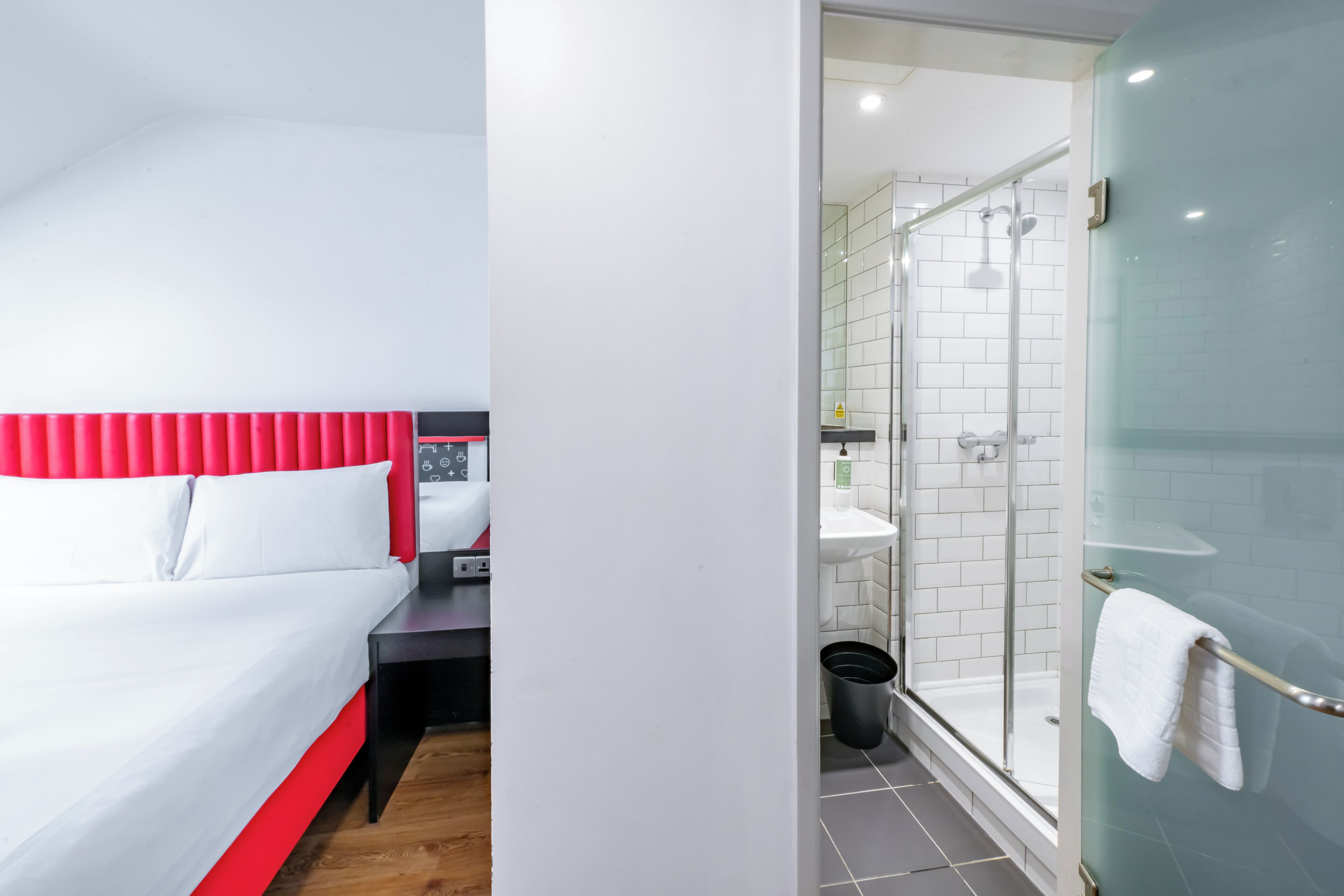 A hotel room with a bed, a nightstand, a bathroom with a glass shower enclosure, and a white towel hanging on the shower door.