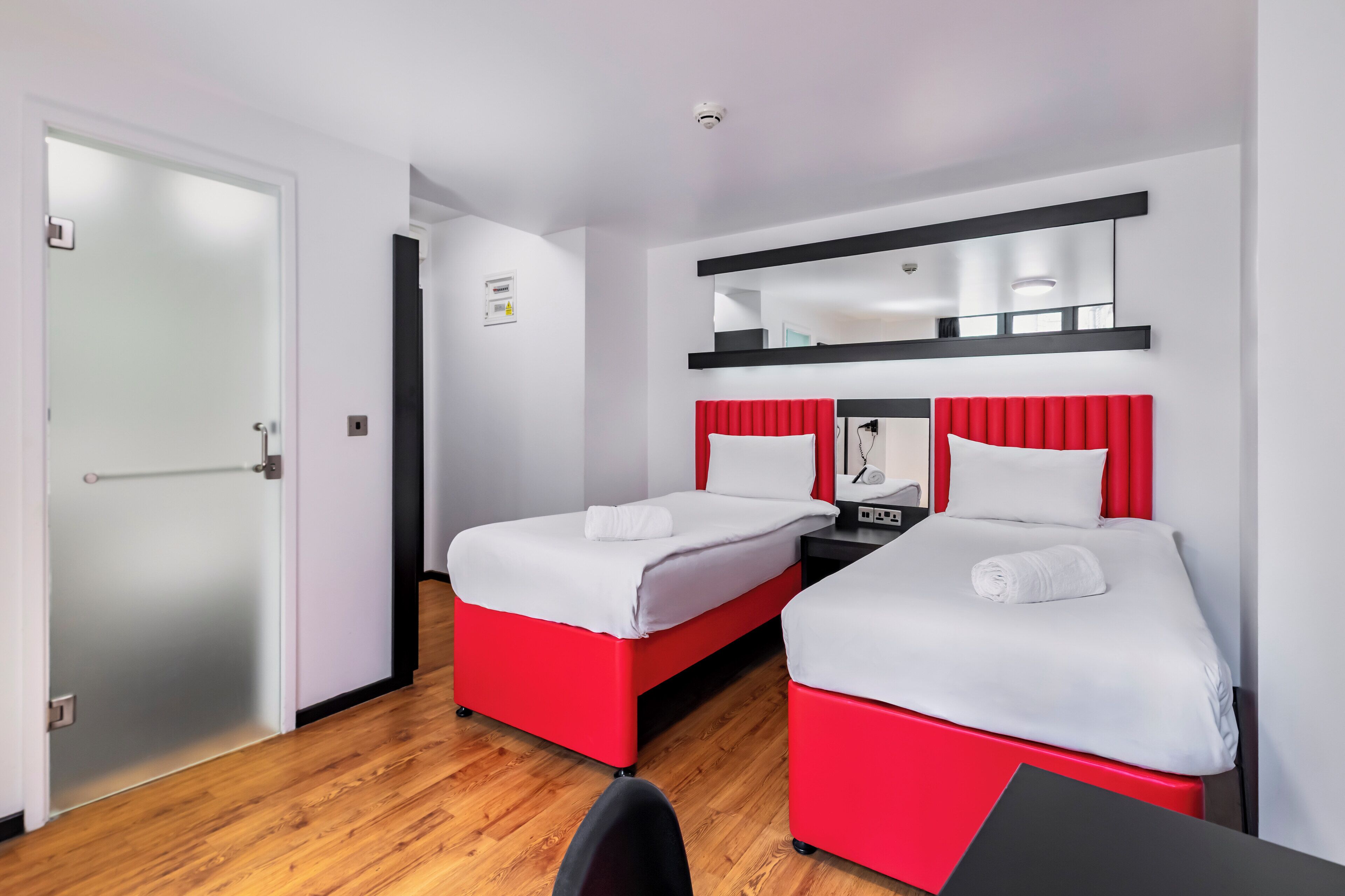 A hotel room with two beds, each with red headboards and white bedding. There is a desk with a chair, a small table, and a mirror above the bed. The room has white walls and a wooden floor.