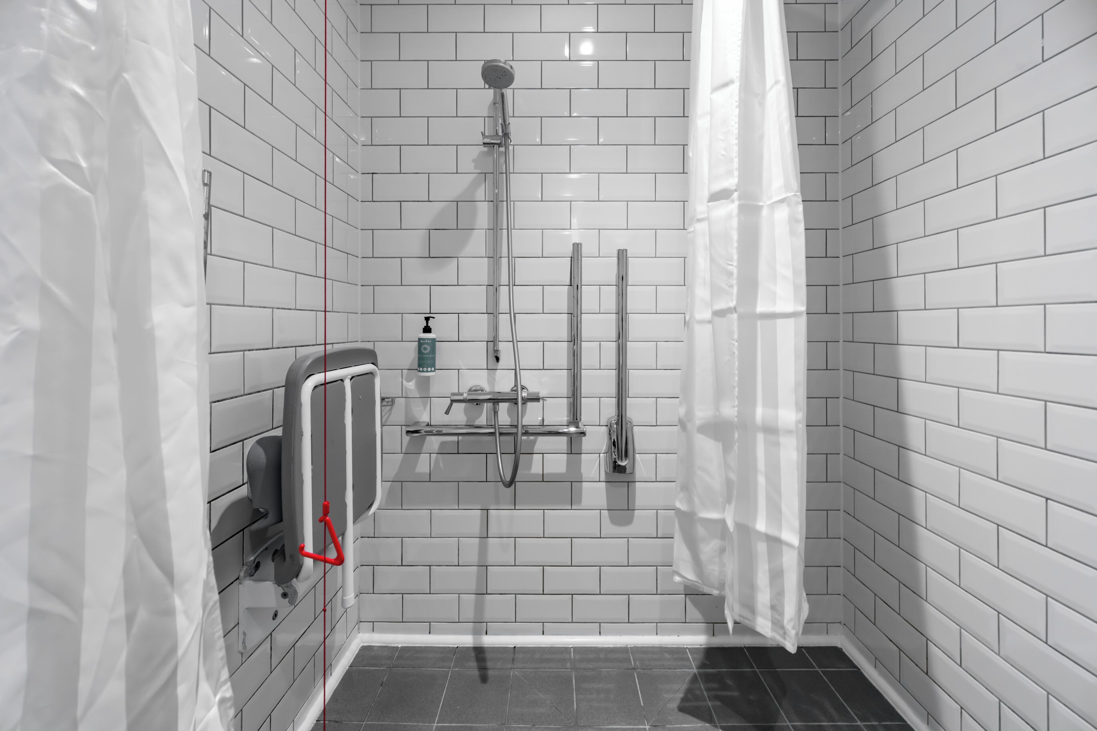 A small, modern bathroom with white tiled walls, a shower with a handheld showerhead, a handheld showerhead, and a soap dispenser.