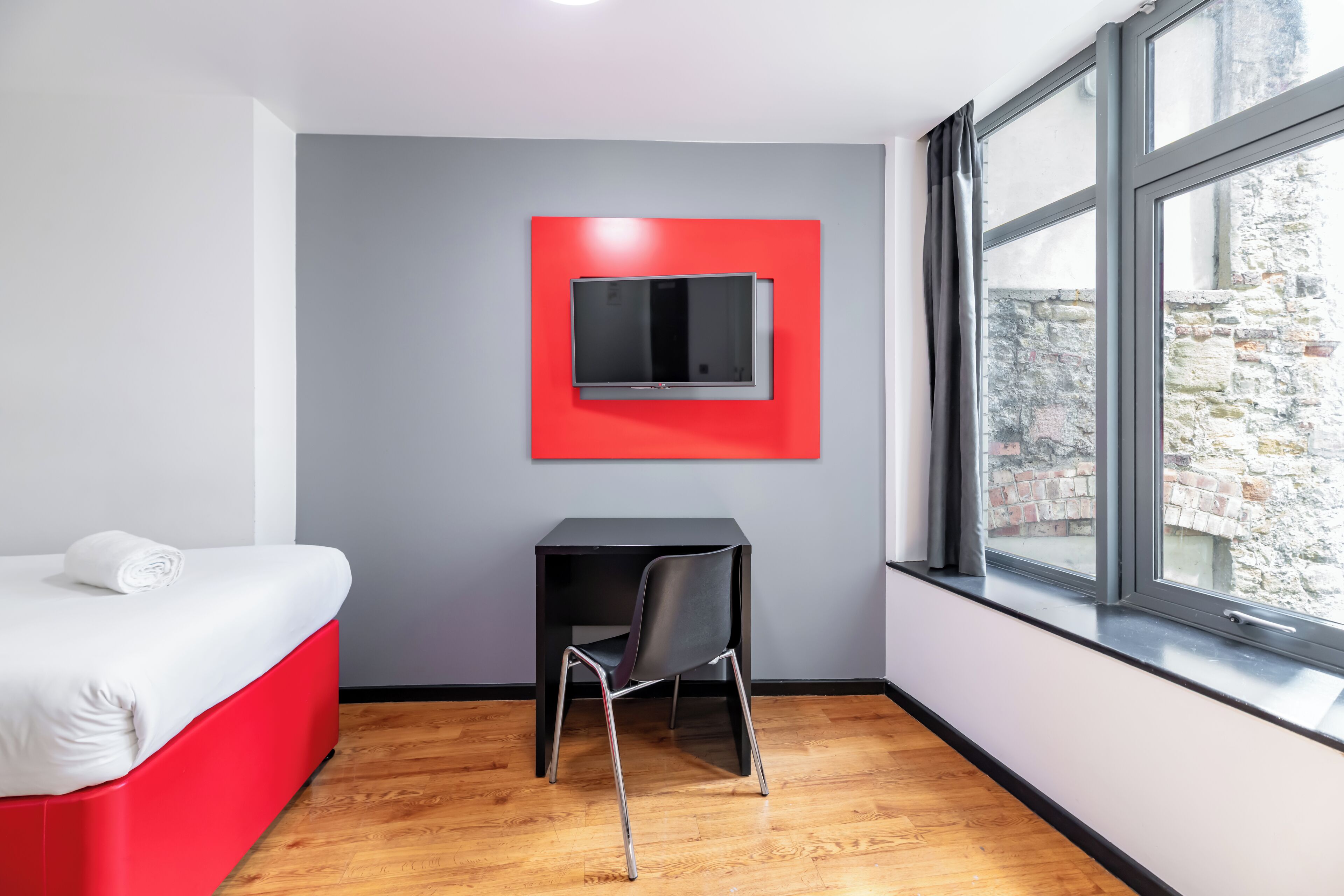 A modern room with a bed, a desk, and a TV mounted on a red panel.