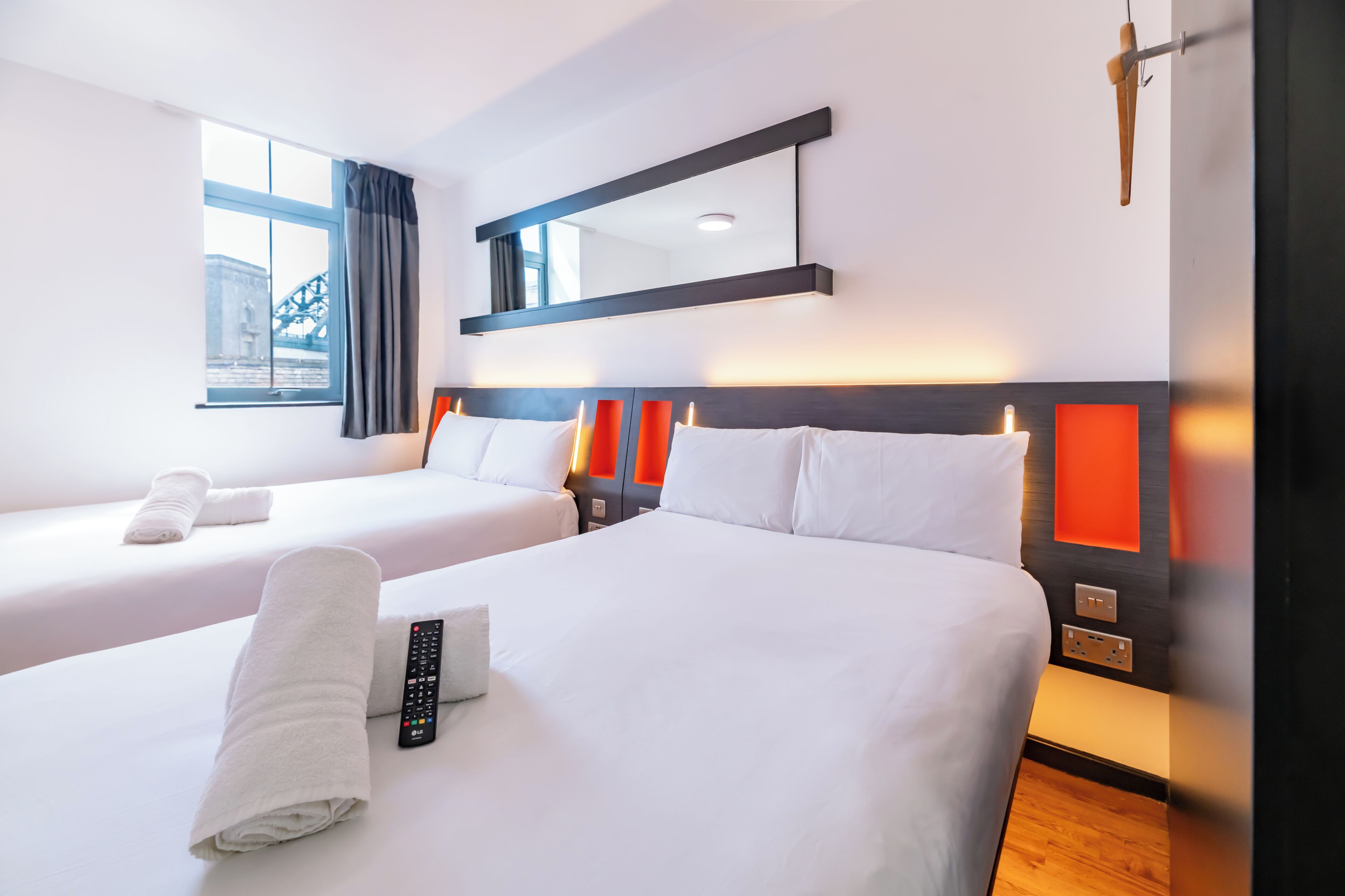 A hotel room with two beds, a large window with curtains, a mirror, and a bedside table with a remote control.