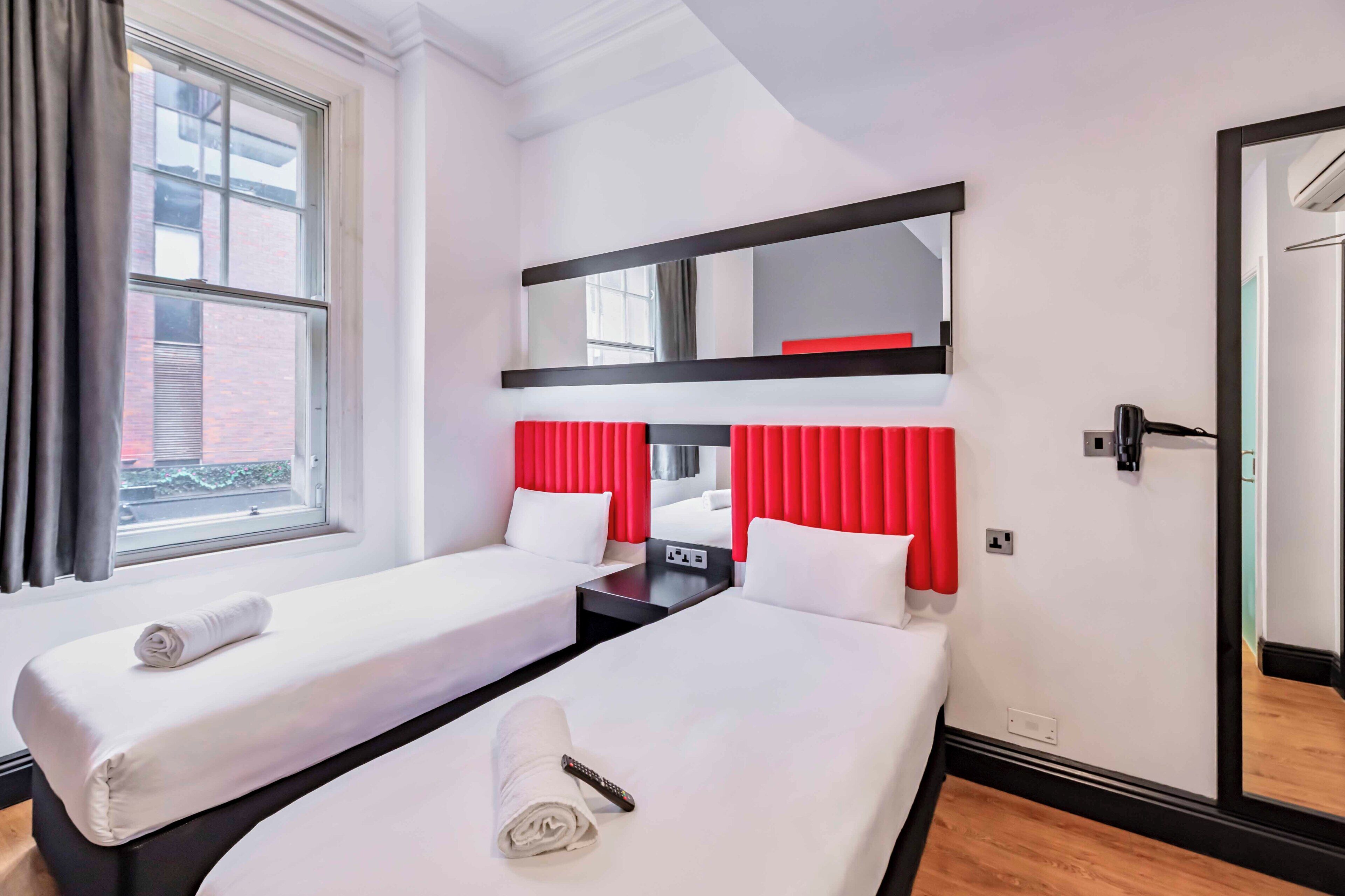 A hotel room with two beds, a mirror, a desk with a lamp, and a window.