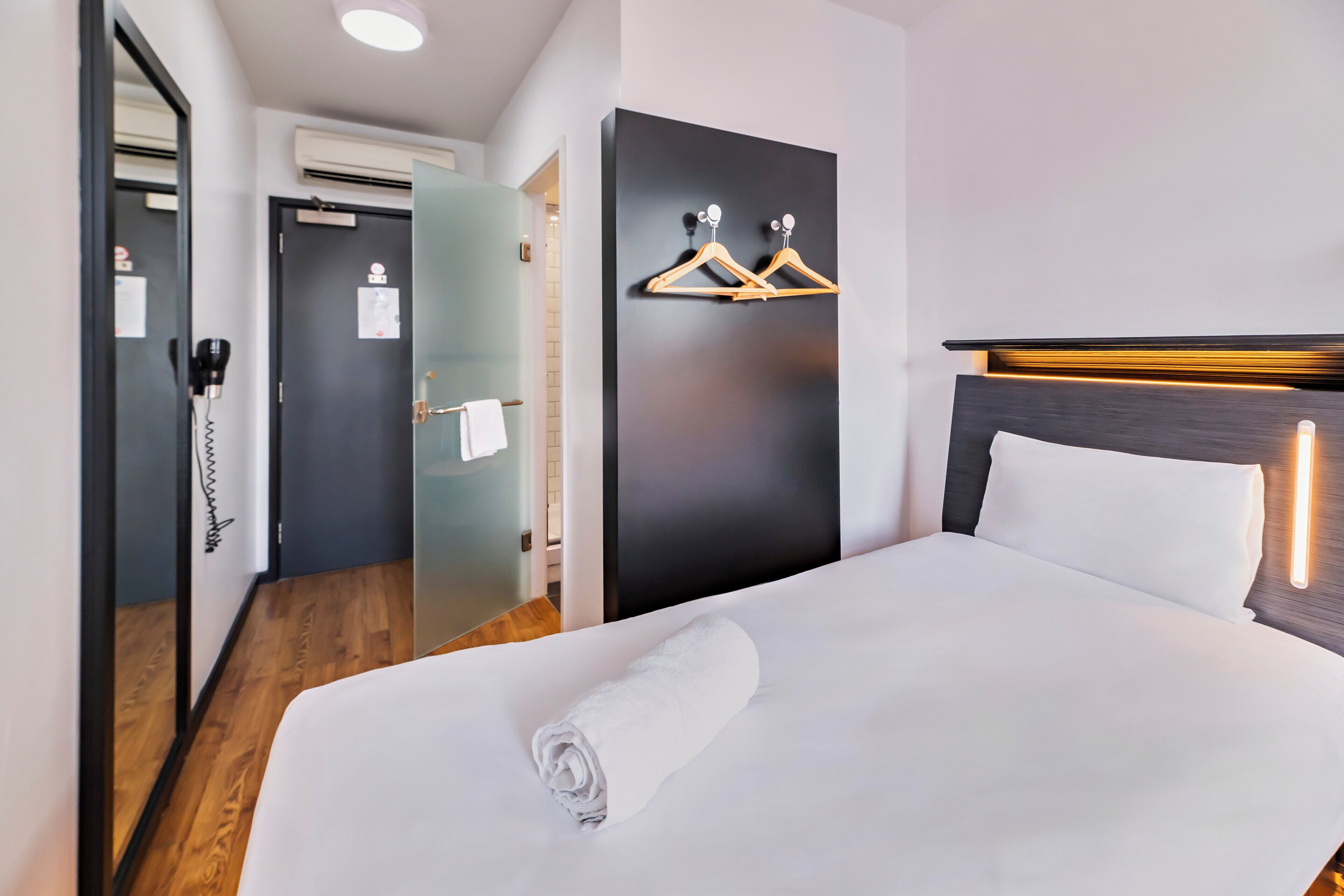 Habitación de hotel con una cama bien hecha, piso de madera, pared negra con una lámpara y una puerta con una toalla colgada.
